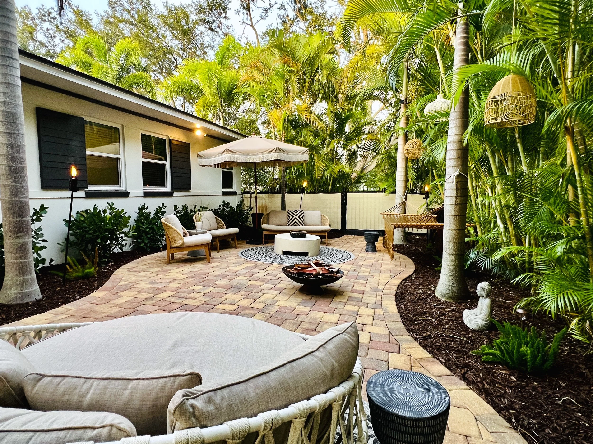 A backyard patio with wicker furniture, an umbrella, a fire pit, and lush tropical plants, including palm trees and hanging wicker lanterns.