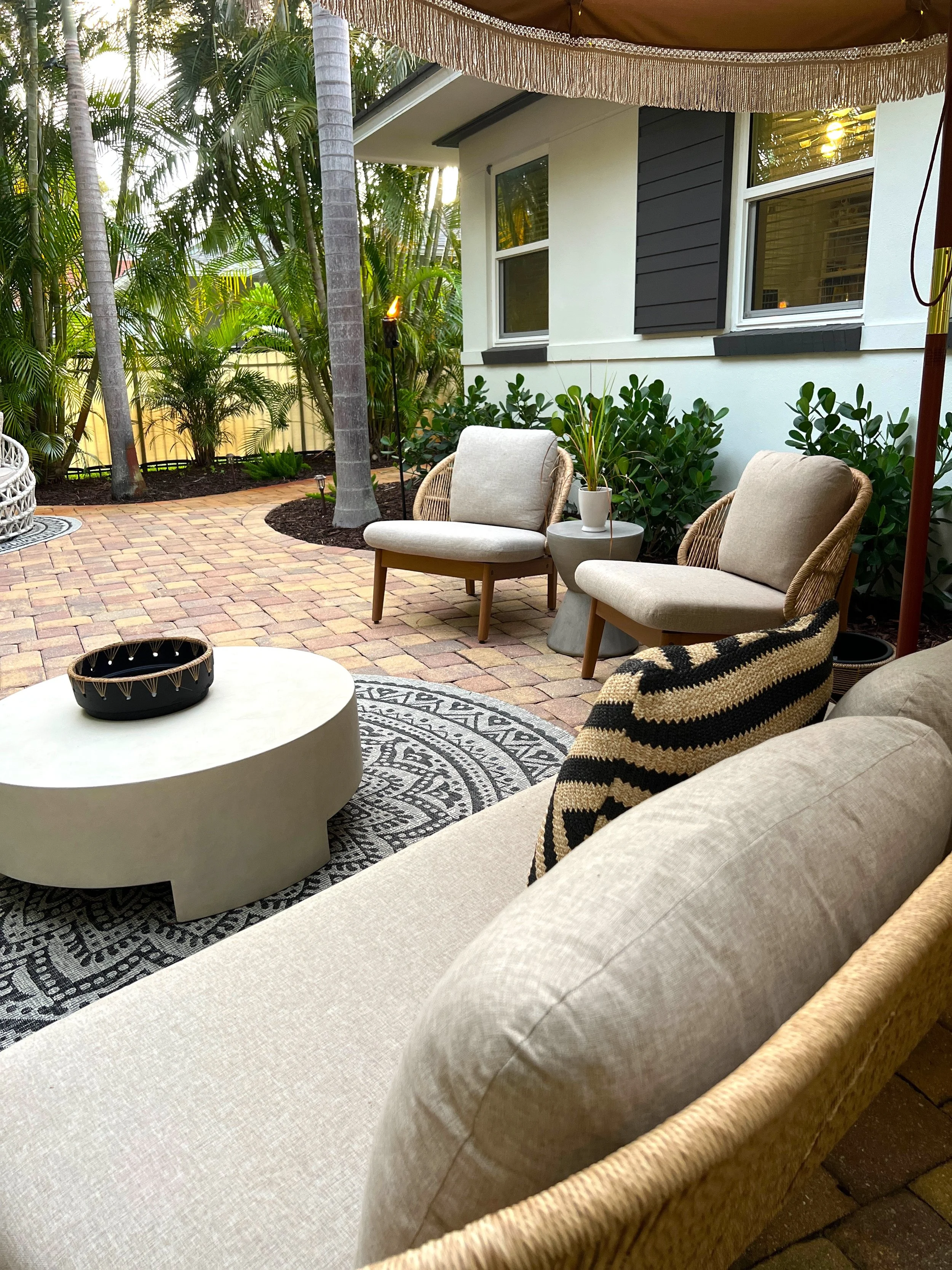 Outdoor patio with wicker chairs, a beige cushion sofa, potted plants, a round coffee table with a black decorative bowl, a patterned rug, and tropical trees with a house in the background.