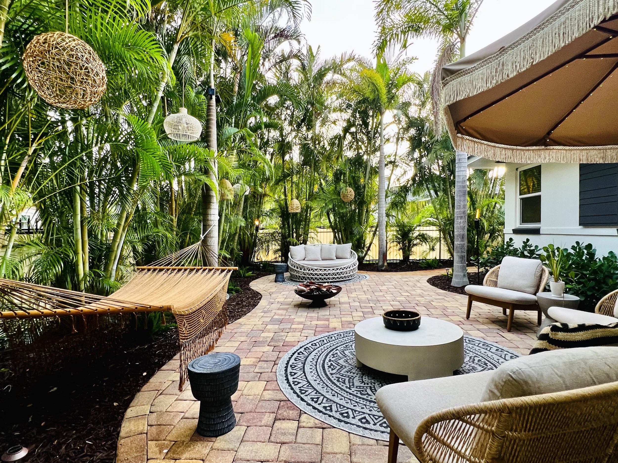 A backyard patio area with wicker furniture, a white circular table, and hanging wicker and fabric lanterns among lush green tropical plants and trees, with part of a house visible.
