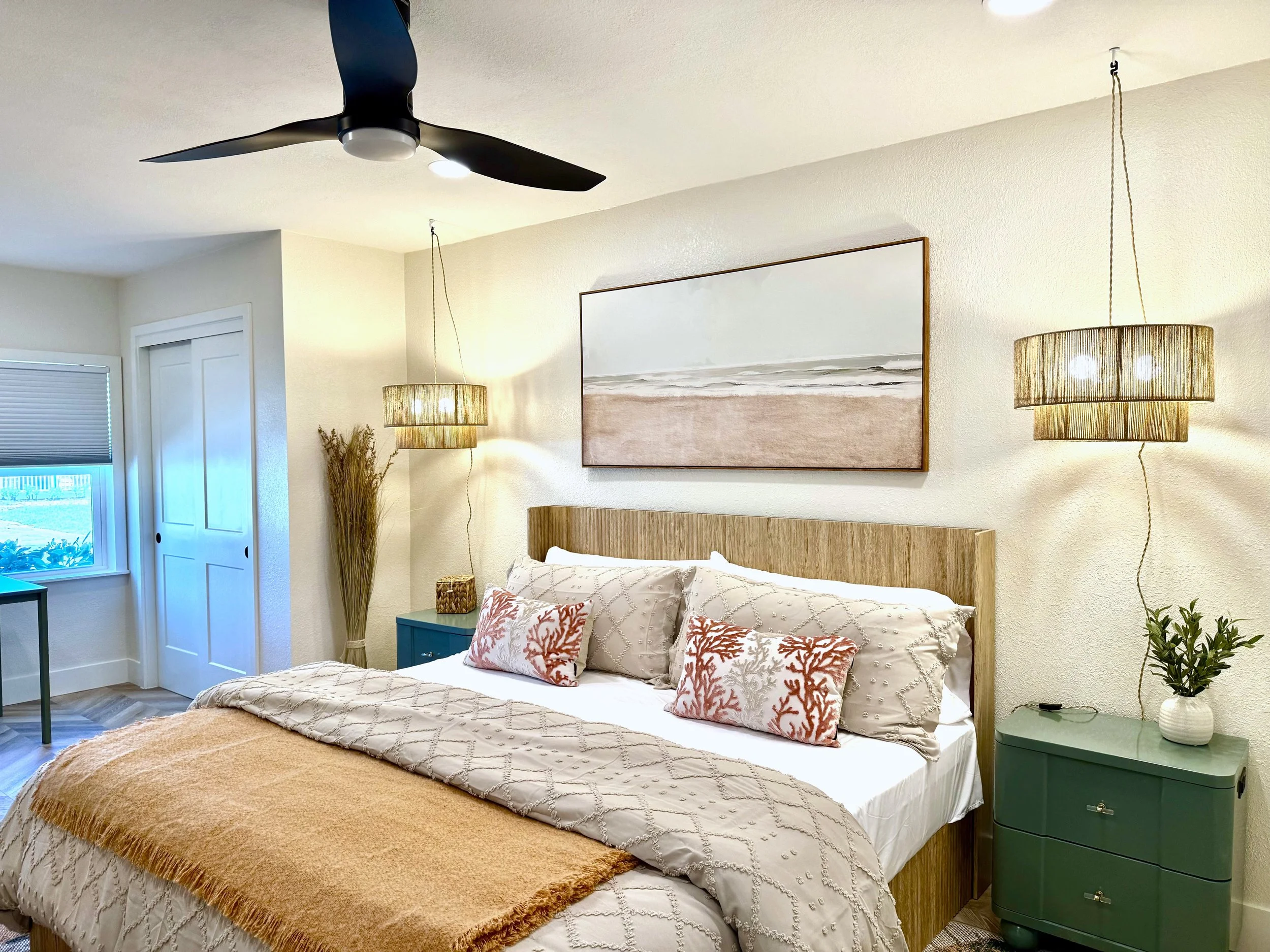 A bedroom with a bed that has white and beige bedding, decorative pillows with coral reef patterns, a wooden headboard, and a tan throw blanket. There are two hanging lamps with beige shades on either side of the bed, a painting of a beach scene abov