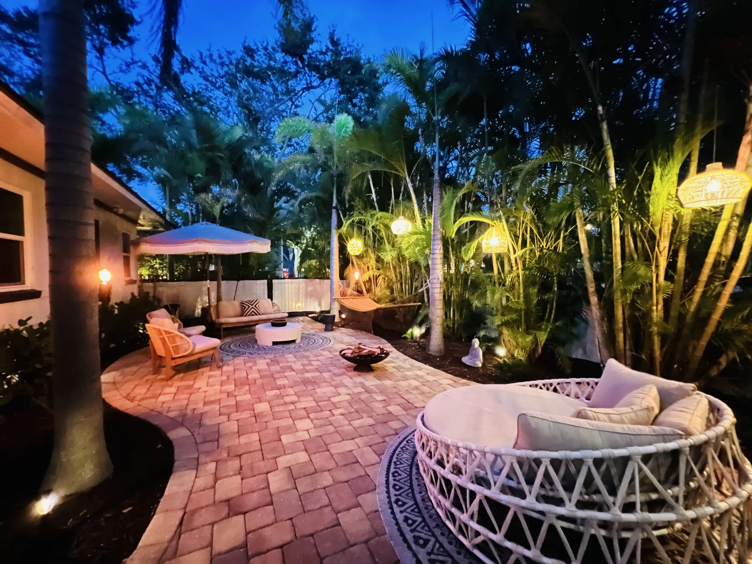 A cozy backyard patio during the evening with outdoor seating, a round white table, a woven lounge chair, and tropical trees illuminated by warm decorative string lights and lanterns.
