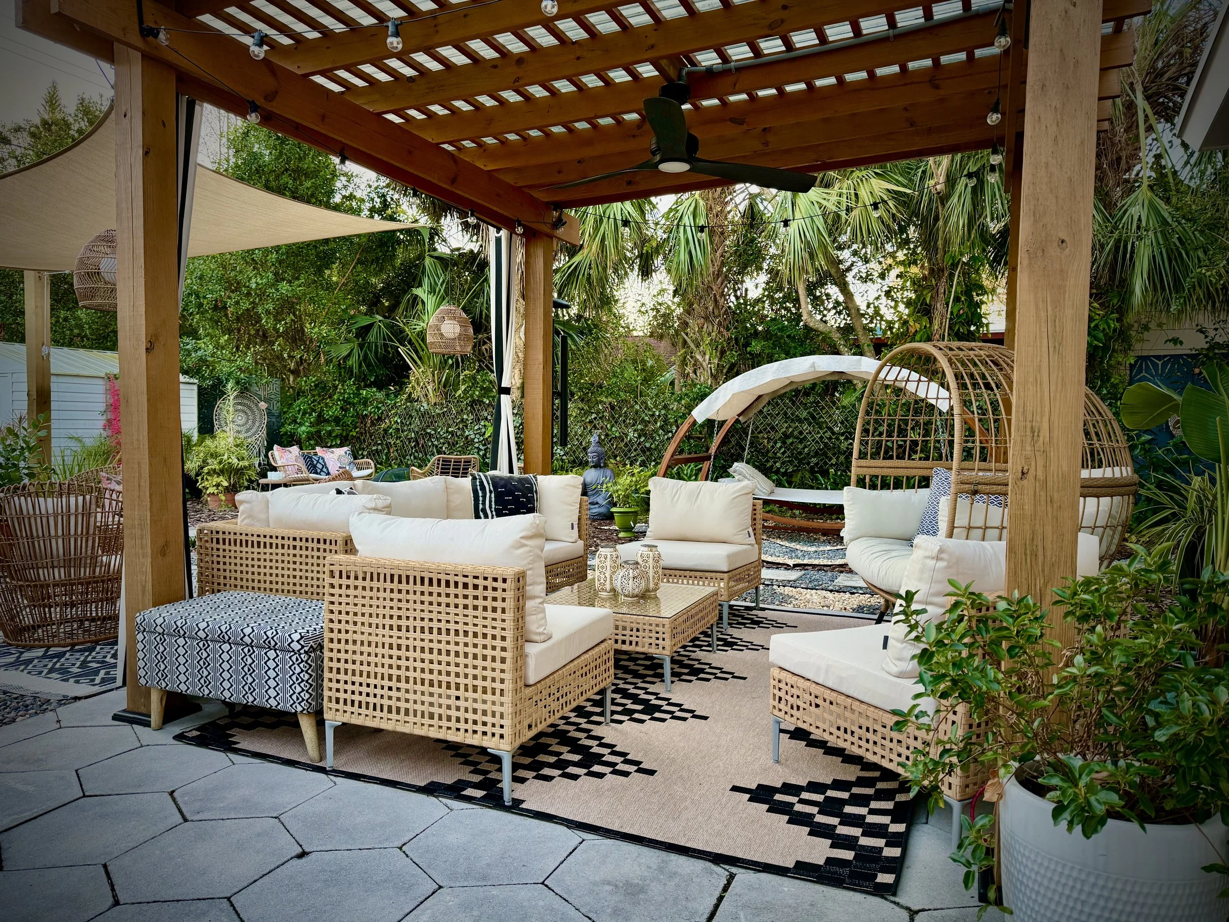 Outdoor patio area with wicker furniture, white cushions, patterned rugs, hanging wicker lights, a black ceiling fan, lush greenery, and a wooden pergola with string lights.