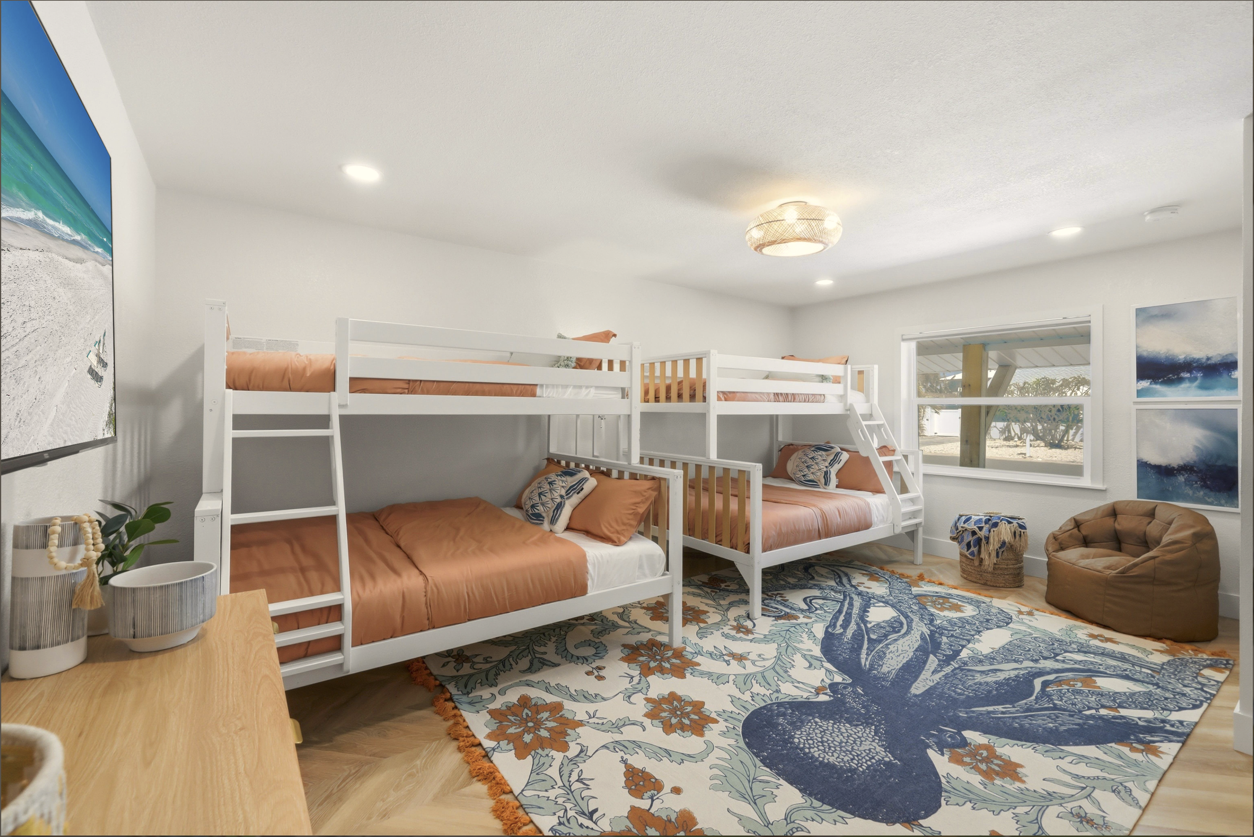Bedroom with white bunk beds with orange bedding, a patterned area rug, and a window showing a beach scene outside.