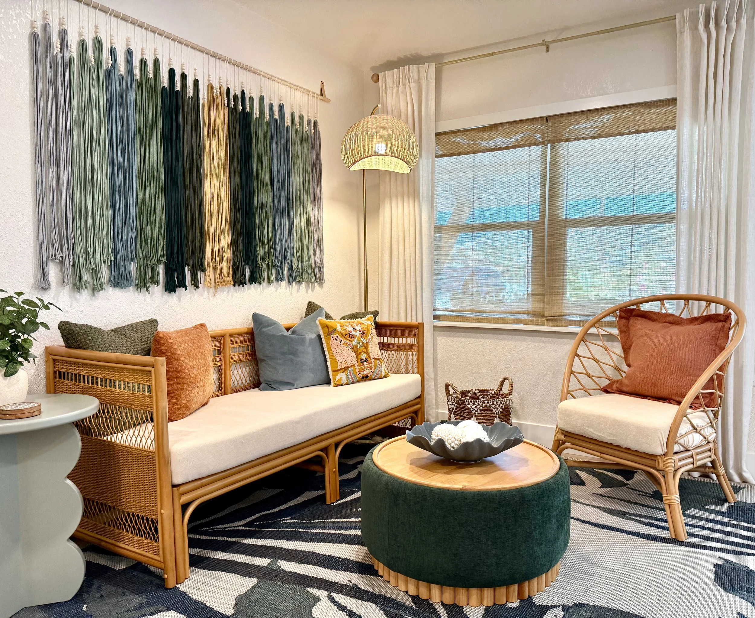 Living room with a woven rattan sofa, orange and blue pillows, a wicker chair, a small round table, a green velvet ottoman, a modern floor lamp, and a wall hanging made of multicolored strings. Window with bamboo shades and sheer white curtains.