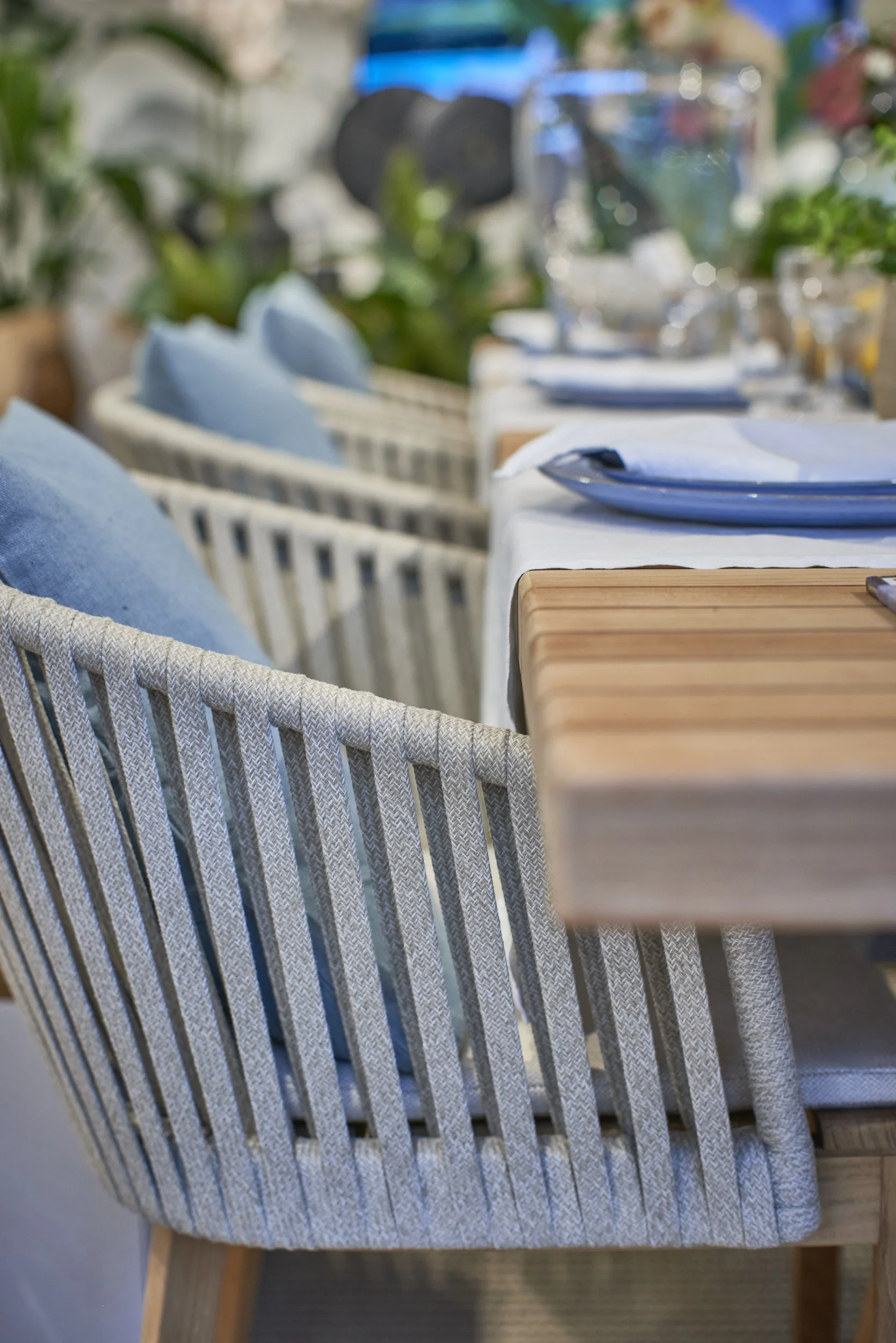 Close-up of a wicker chair with light blue cushions and a wooden table set with a white tablecloth, blue plates, and silverware, in a brightly lit outdoor dining area with potted plants in the background.