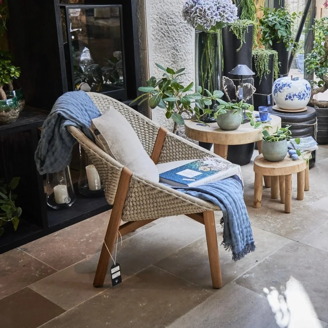 Indoor space with wicker chair draped with a blue blanket and pillow, surrounded by potted plants, a wooden table, and decorative ceramics.