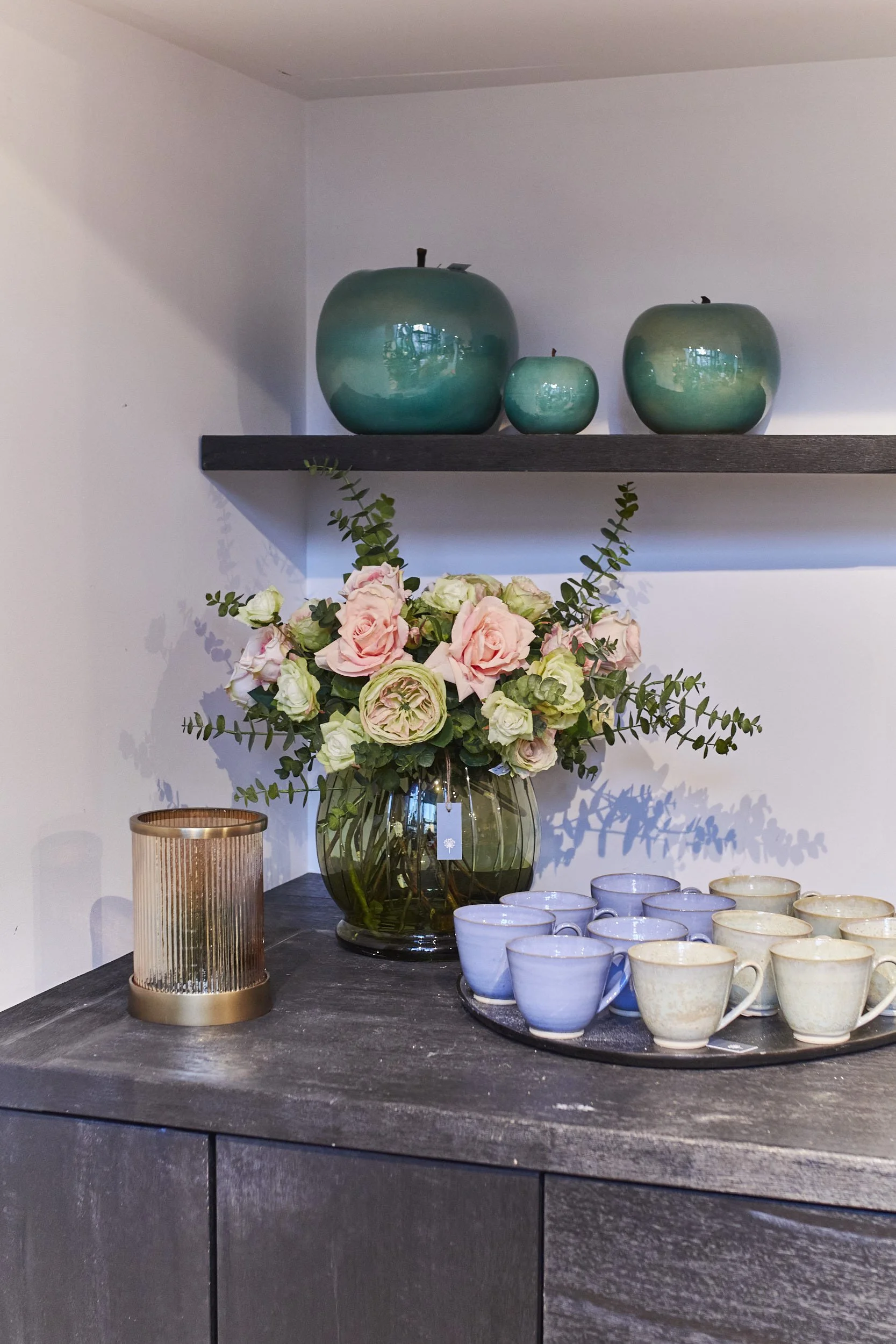 Decorative shelf with green vases, a bouquet of pink and white flowers, a lit candle, and ceramic mugs on a dark countertop.