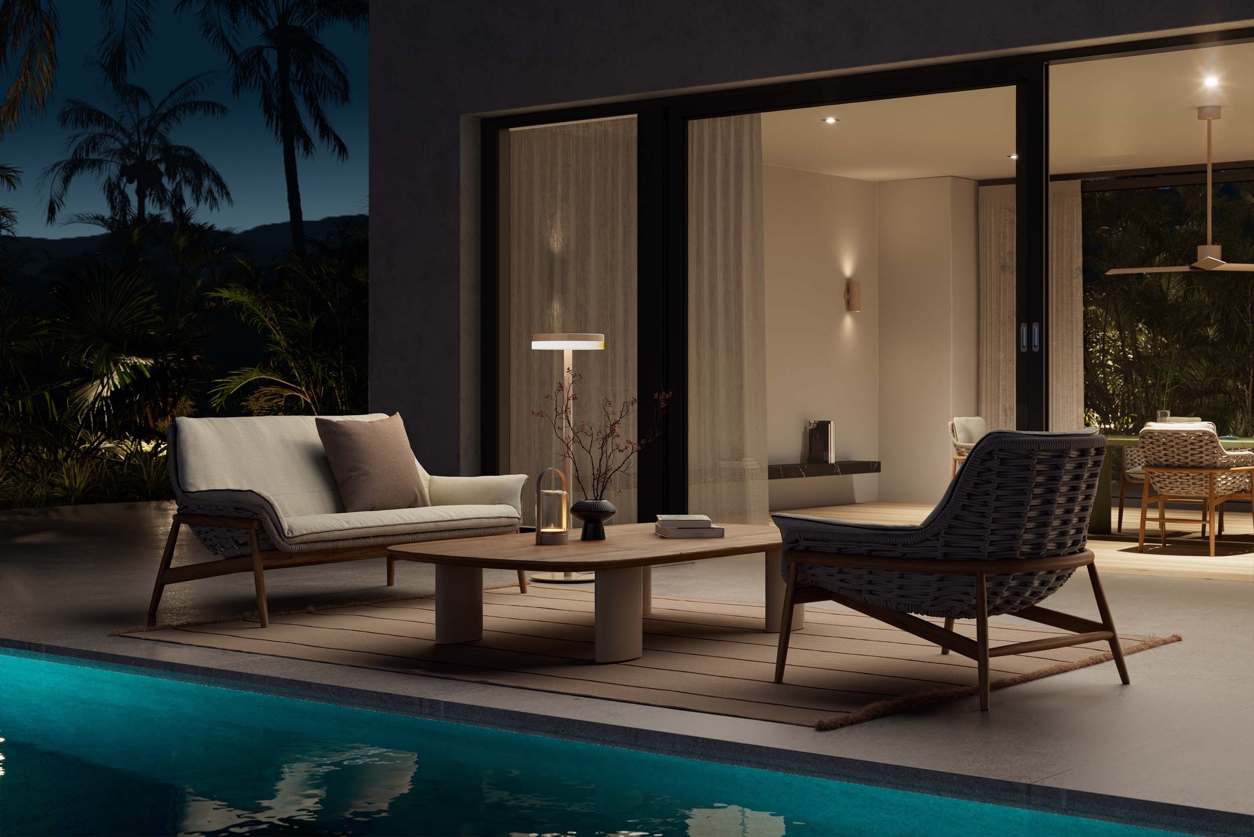 Outdoor patio at night with seating area including a white sofa with a pillow, a wooden chair, a coffee table with a lamp, decorative items, and an indoor dining area visible through glass sliding doors, with lush greenery and palm trees in the background.