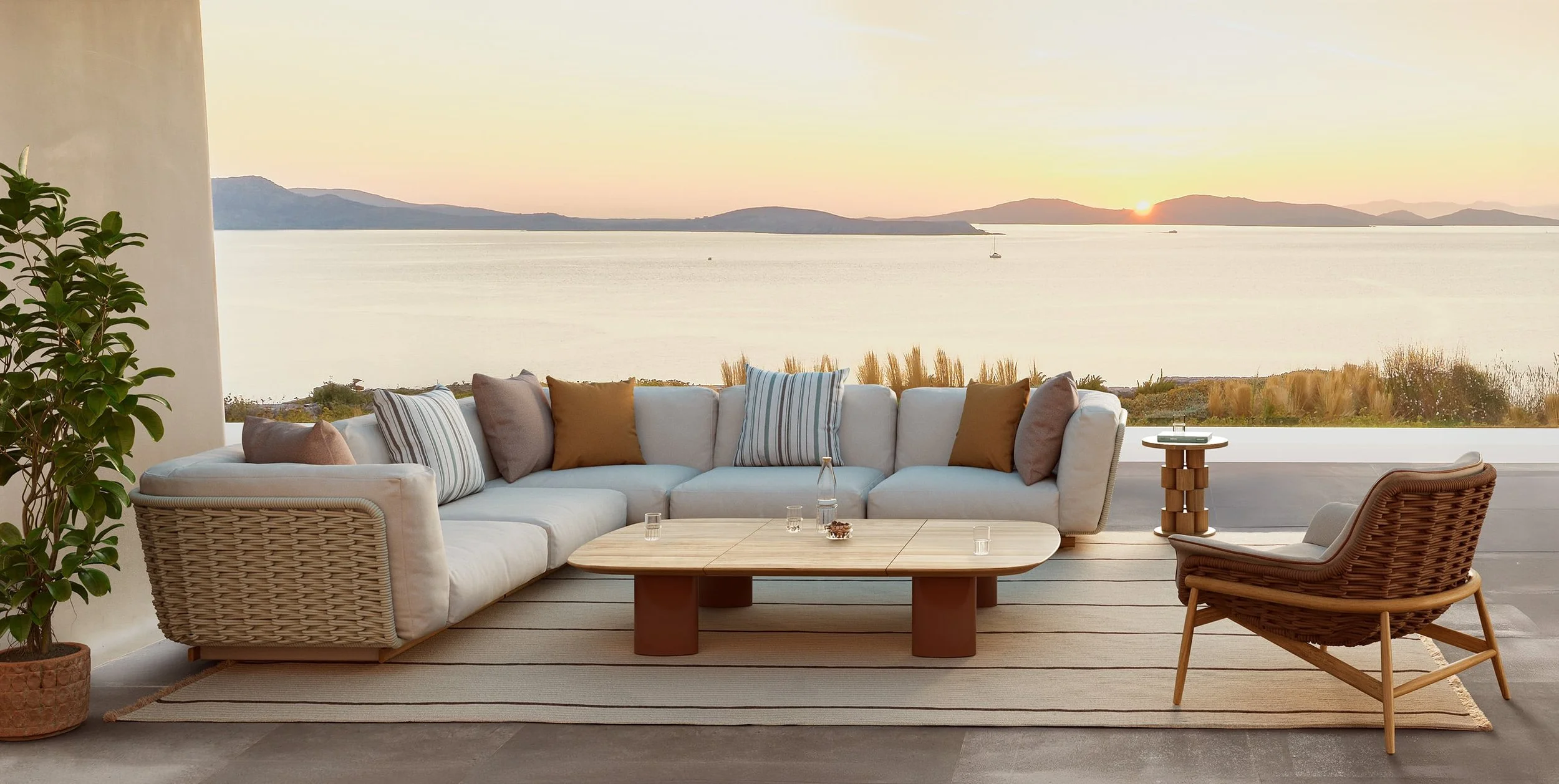 Modern outdoor living room with a large gray sectional sofa, striped and solid throw pillows, a wooden coffee table, a round side table, and a rattan chair, all facing a scenic view of a lake and mountains at sunset.