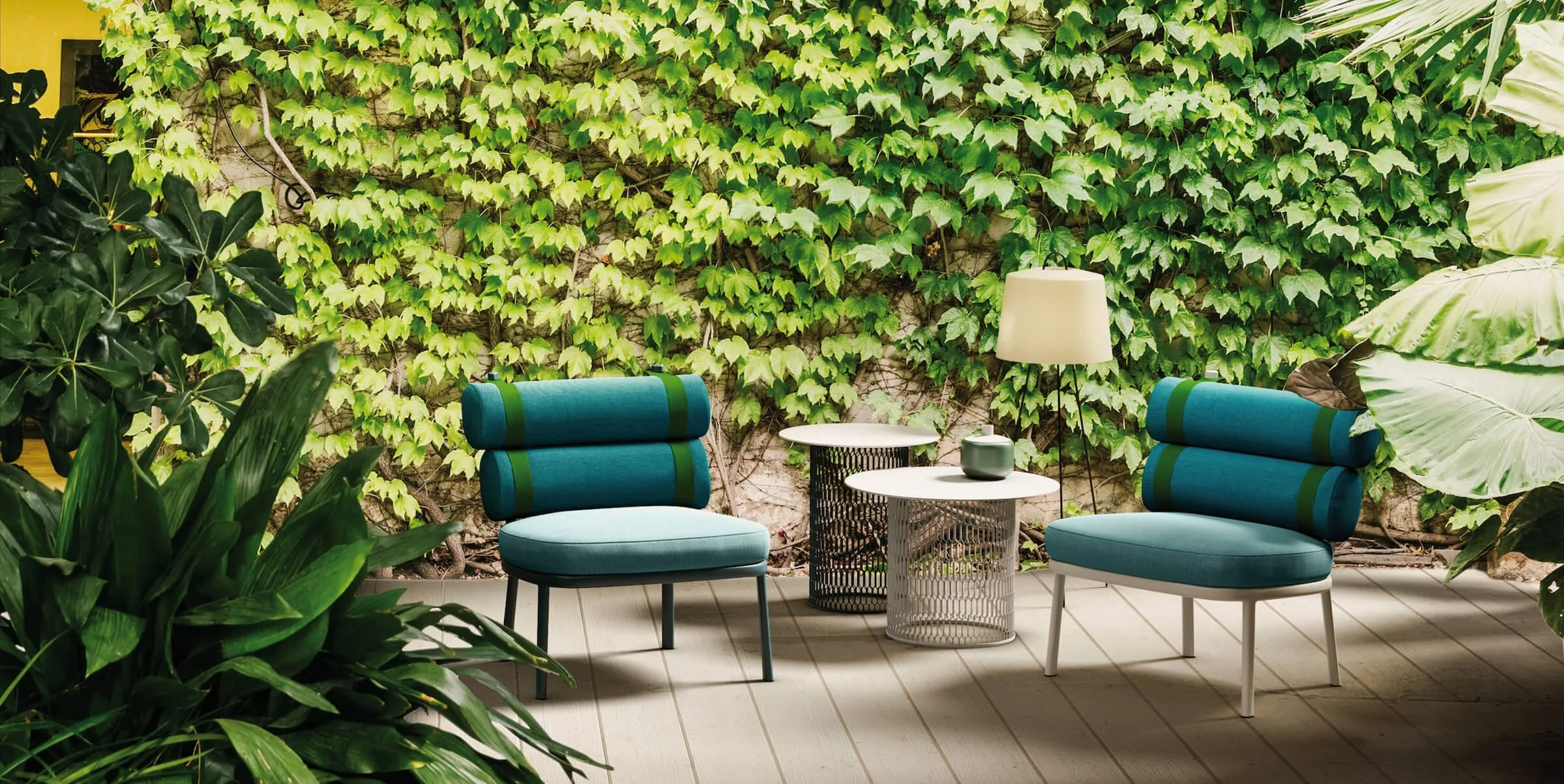 Outdoor seating area with two blue chairs featuring green stripes, a round white table, a side table with a wireframe base, an off-white lamp, and surrounding lush green plants and ivy-covered wall.