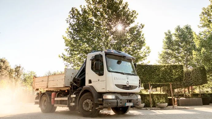 A white flatbed truck parked outdoors among trees and landscaped shrubs, with sunlight shining through the trees.