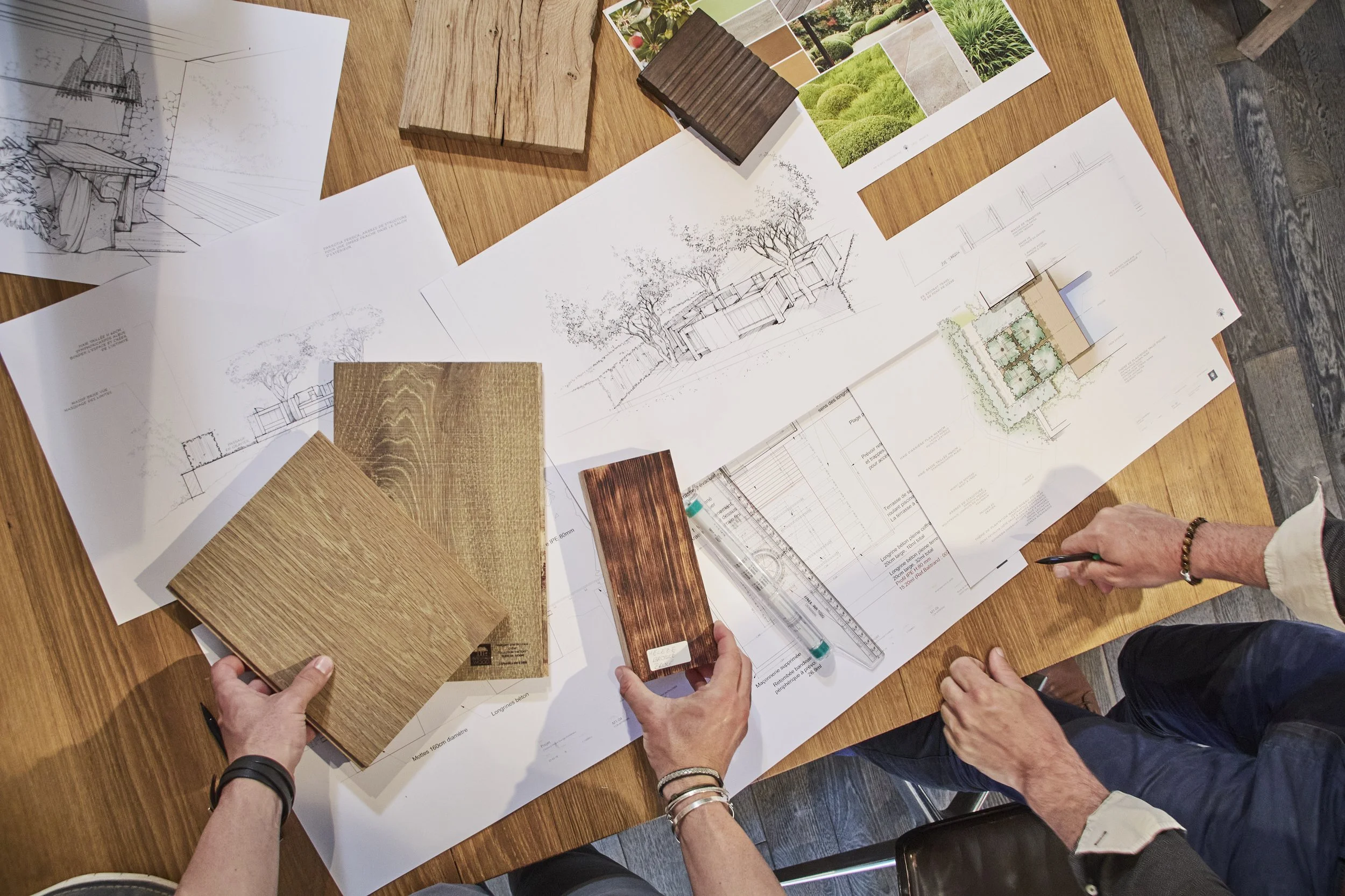 A top view of a wooden table with architectural drawings, photo samples of landscaping, and color swatches. Hands of multiple people are visible, some holding pens or color samples, indicating a collaborative planning or design session.