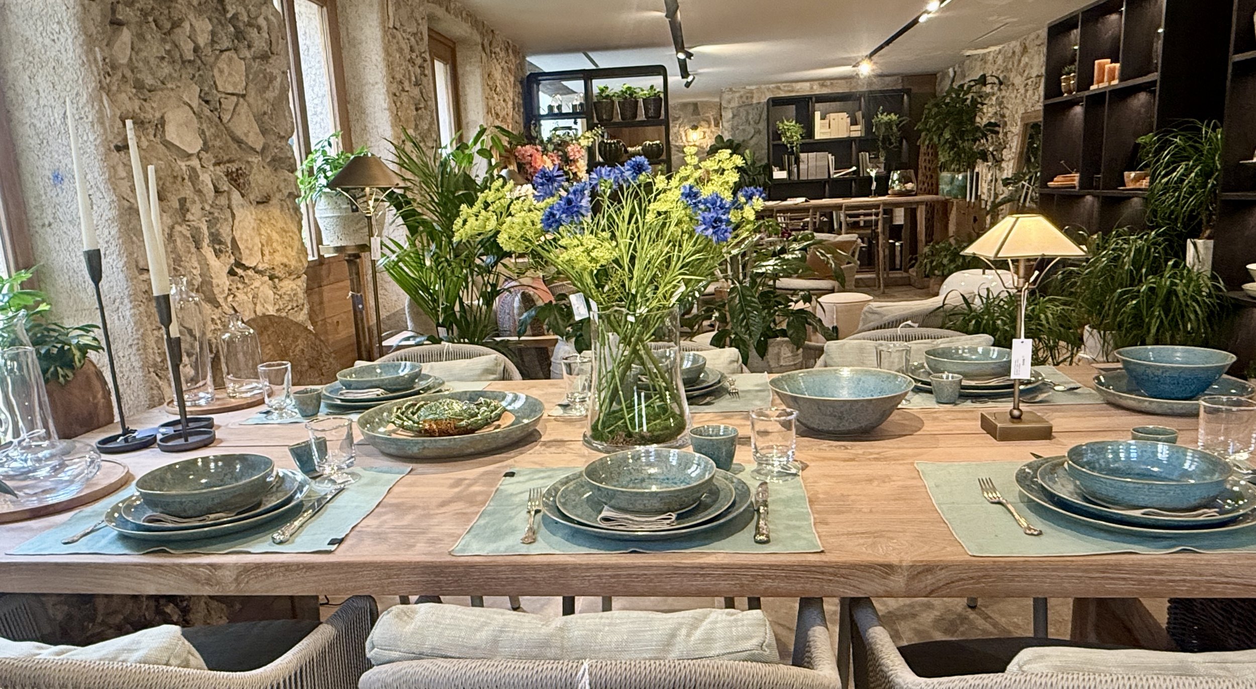 Interior of a rustic dining room decorated with stone walls, lush green plants, a large wooden dining table set with blue and green ceramic dishes, glassware, and candles, illuminated by natural light from windows and warm lamps.