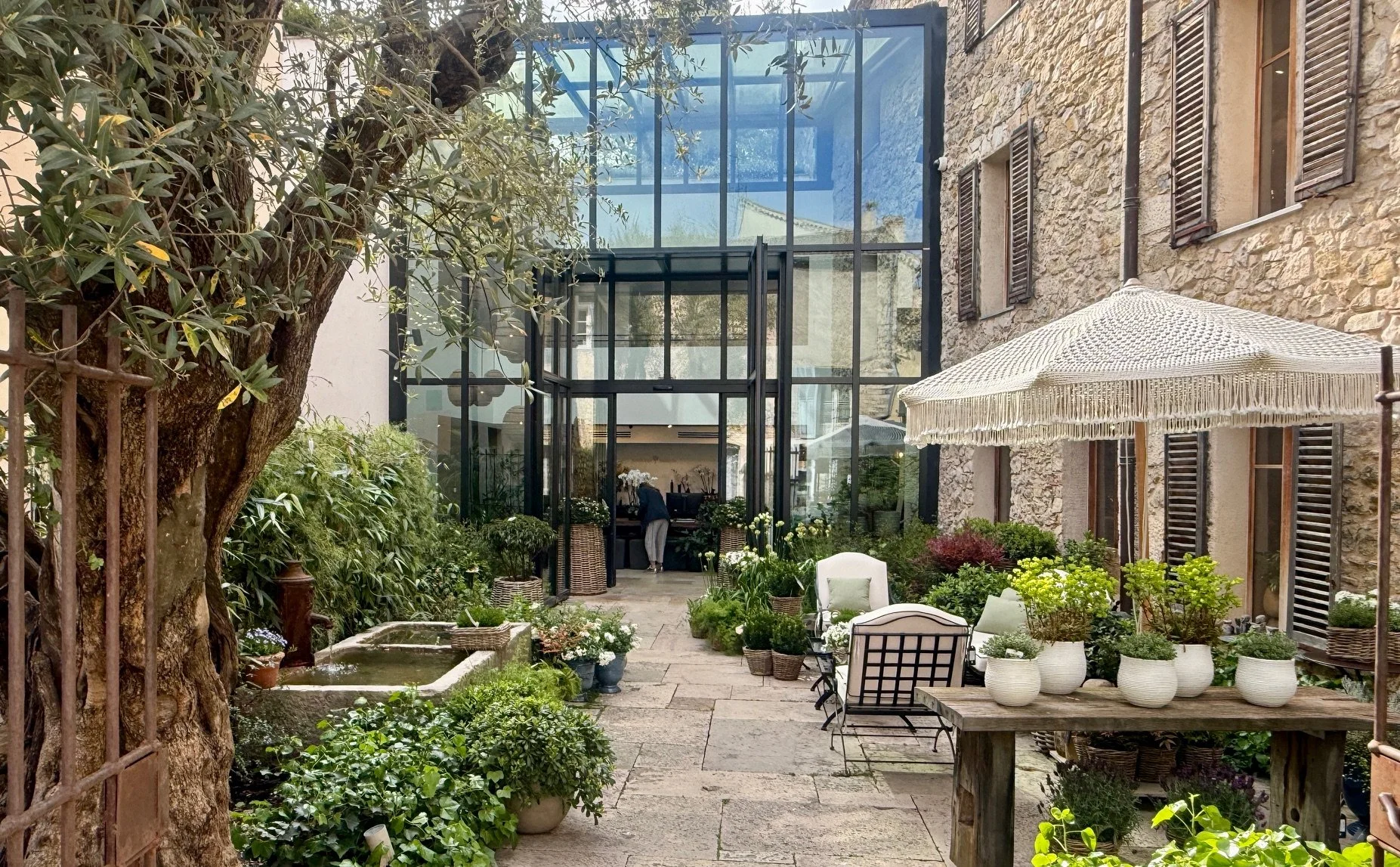 View into the front entrance of the garden boutique, qith the large atrium in the background and the olive tree and stone floored courtyard in the foreground with wrought iron garden furniture and shade umbrella