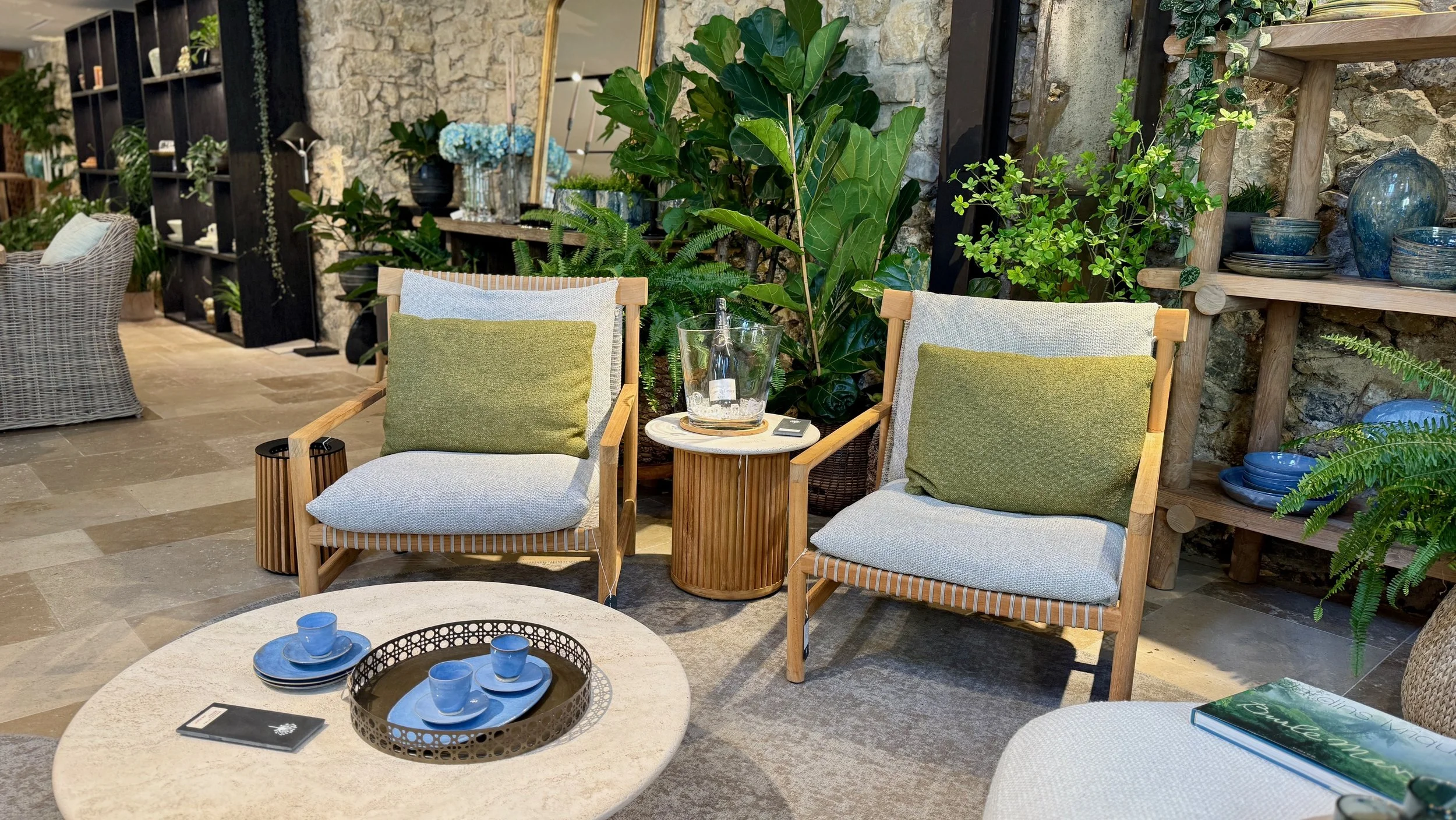 Indoor seating area with wicker and wooden chairs, a round table with blue cups, and a backdrop of green plants, a stone wall, and wooden shelves with decorative ceramics.