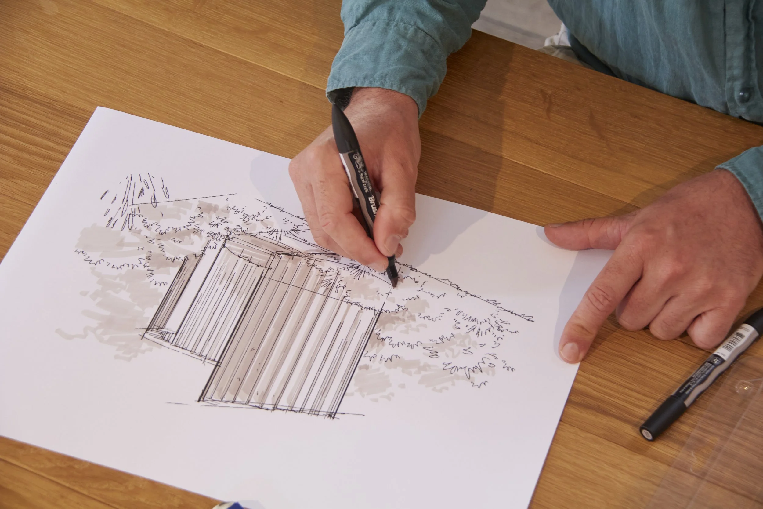 A person drawing an architectural sketch of a house on a white sheet of paper, with some notes around the sketch, while sitting at a wooden table.