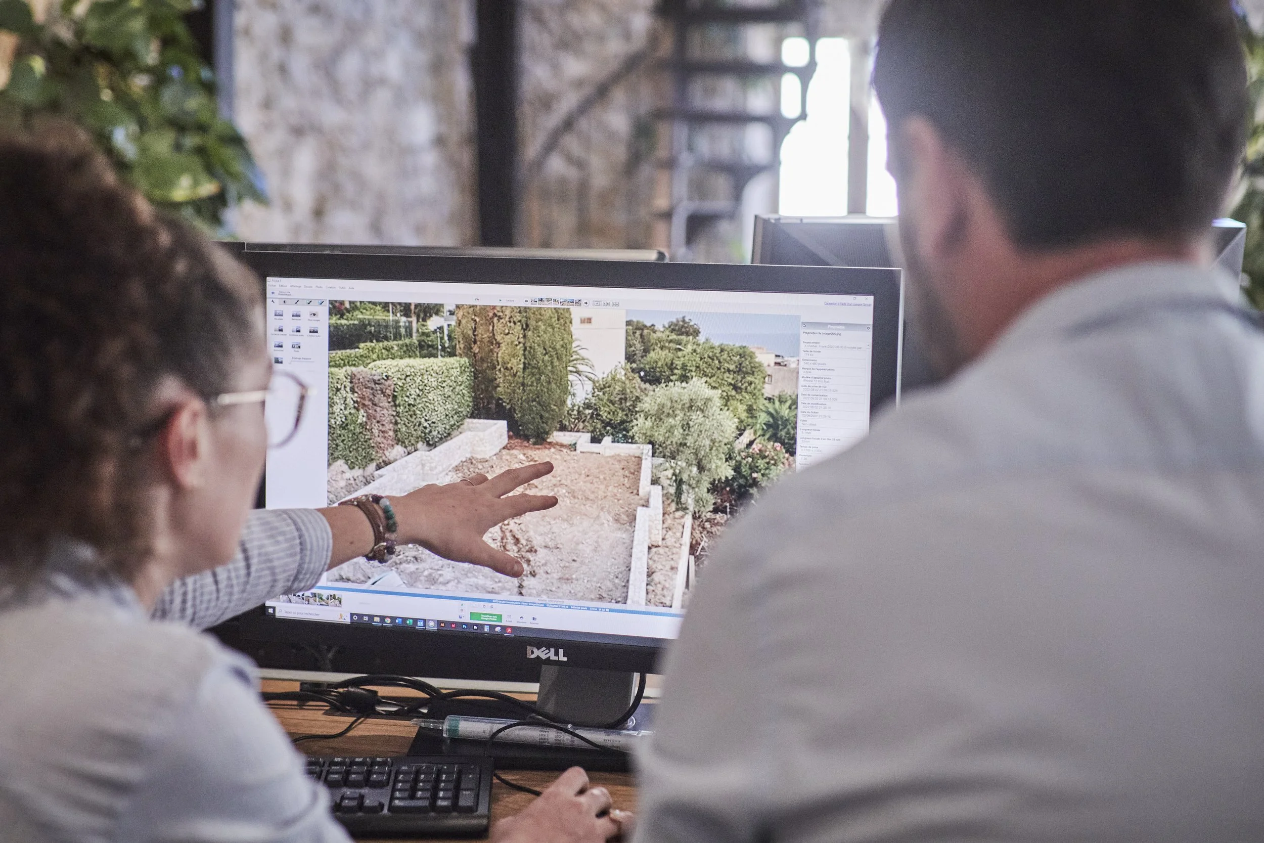 Two people working together at a desk, looking at a computer monitor displaying a garden design or landscape plan with bushes, trees, and a pathway.
