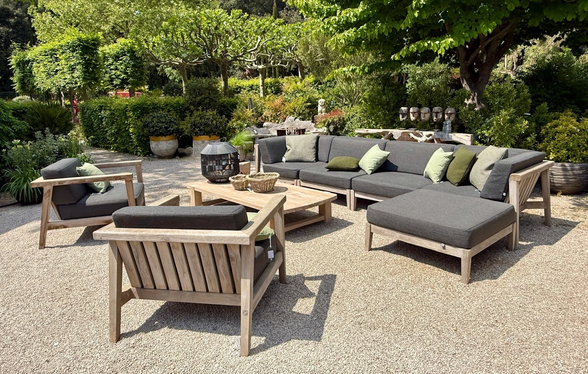 Outdoor seating area with modern wooden furniture and gray cushions surrounded by lush greenery and trees.