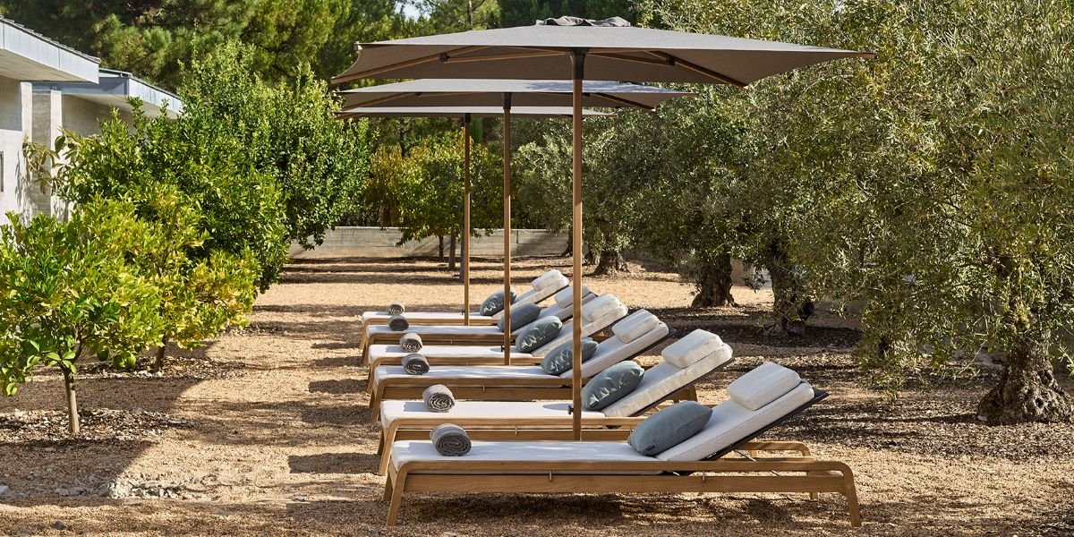 Six wooden lounge chairs with rolled towels and cushions, shaded by large umbrellas, set on a dirt path among trees.