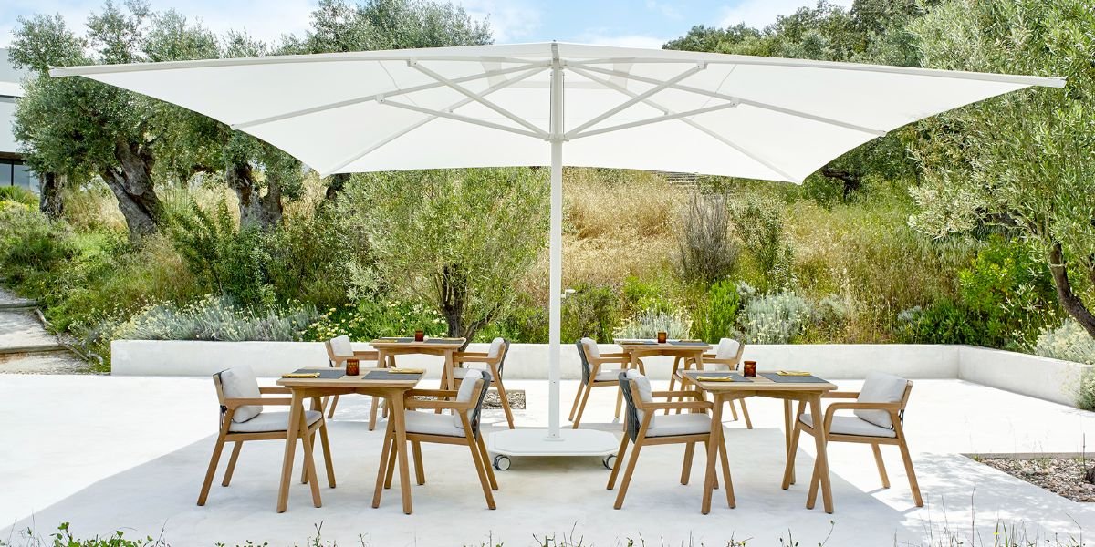 Outdoor patio with six wooden dining tables and chairs under a large white umbrella, surrounded by greenery and trees.