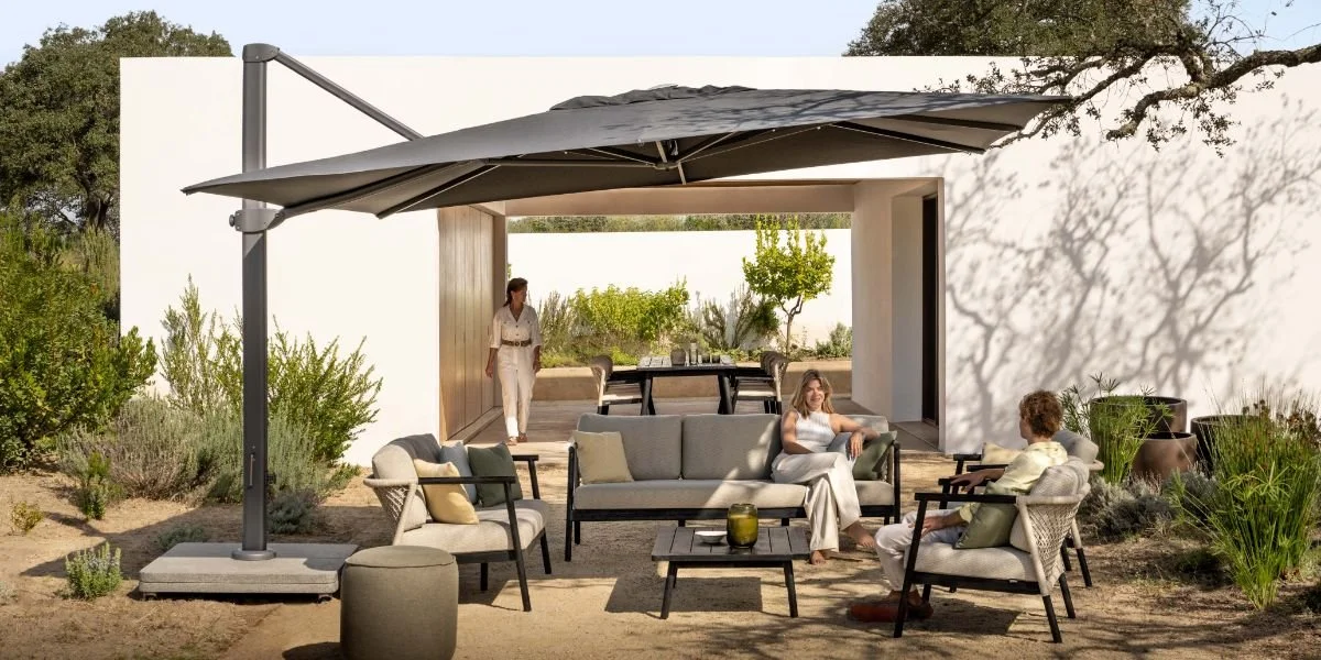 Outdoor patio with modern furniture, large umbrella, and three people sitting and talking, with a person walking in the background.