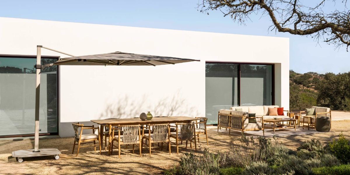 Modern outdoor patio with wooden dining table, chairs, and a seating area with sofa and armchairs, on a dirt ground with desert plants, adjacent to a white building with large glass sliding doors, under a blue sky.