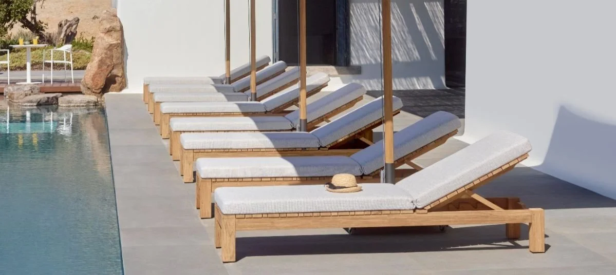 Row of wooden poolside lounge chairs with white cushions, beside a swimming pool, with a straw hat resting on one of the chairs.