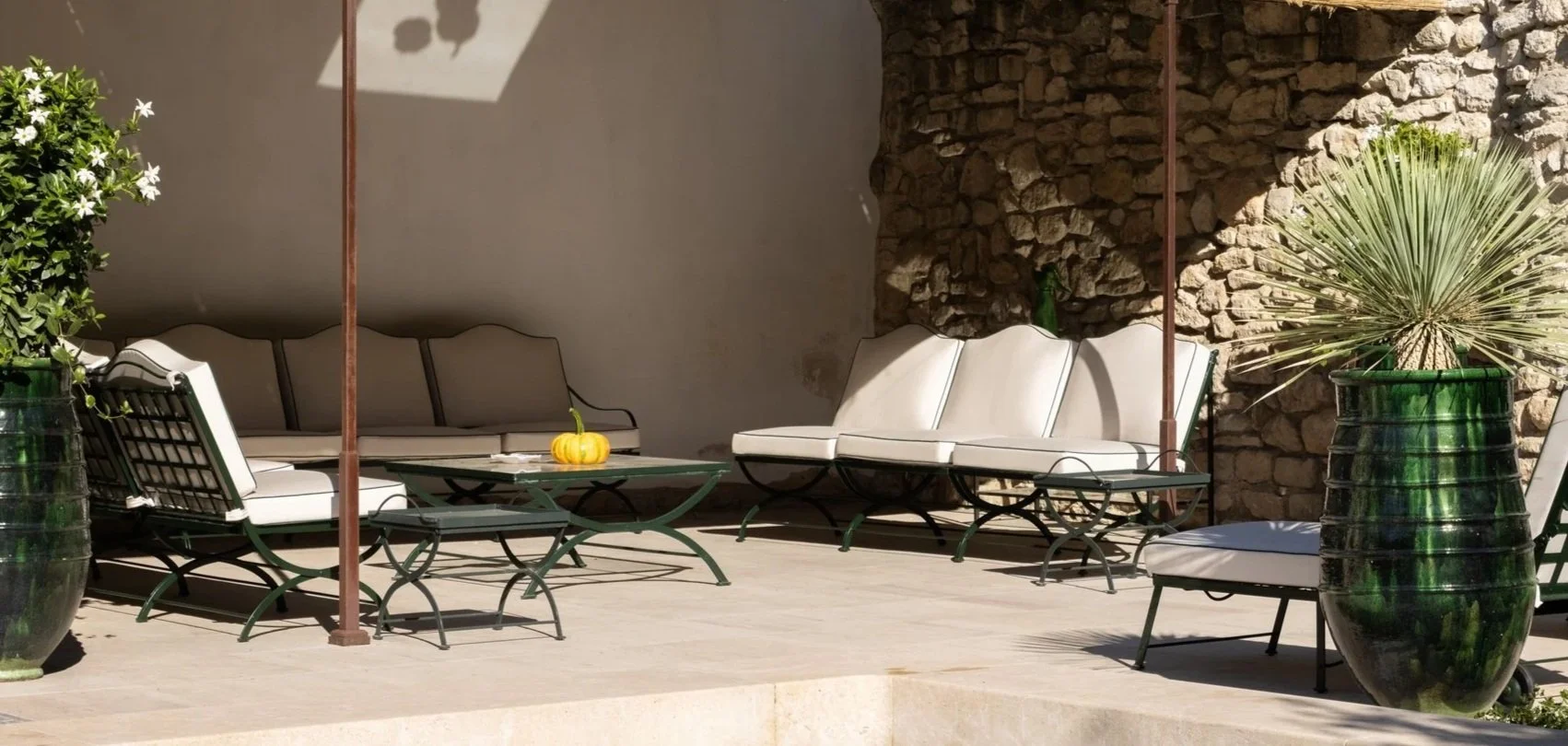 Outdoor patio area with two white cushioned sofas, metal chairs, and a glass table with a small pumpkin. Large green plants in tall glass pots are on either side. A stone wall provides a rustic backdrop, and sunlight casts shadows on the wall.