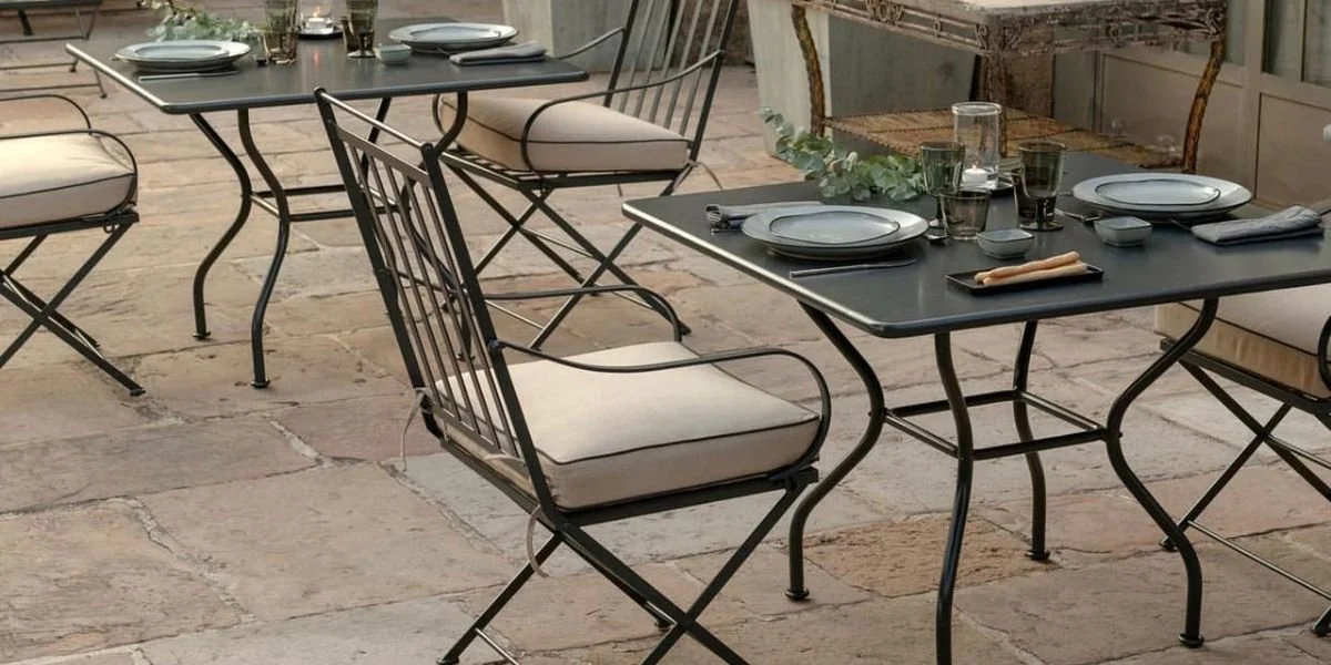 Elegant outdoor dining area with black metal tables and chairs on a stone patio, set with plates, glasses, and a small plant centerpiece.