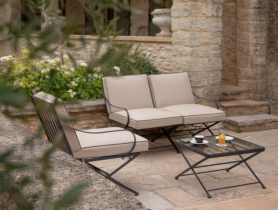 Outdoor patio scene with two beige cushioned chairs, a matching loveseat, and a glass-top coffee table with coffee, orange juice, and cups on it, set against a stone wall with steps and flowers.