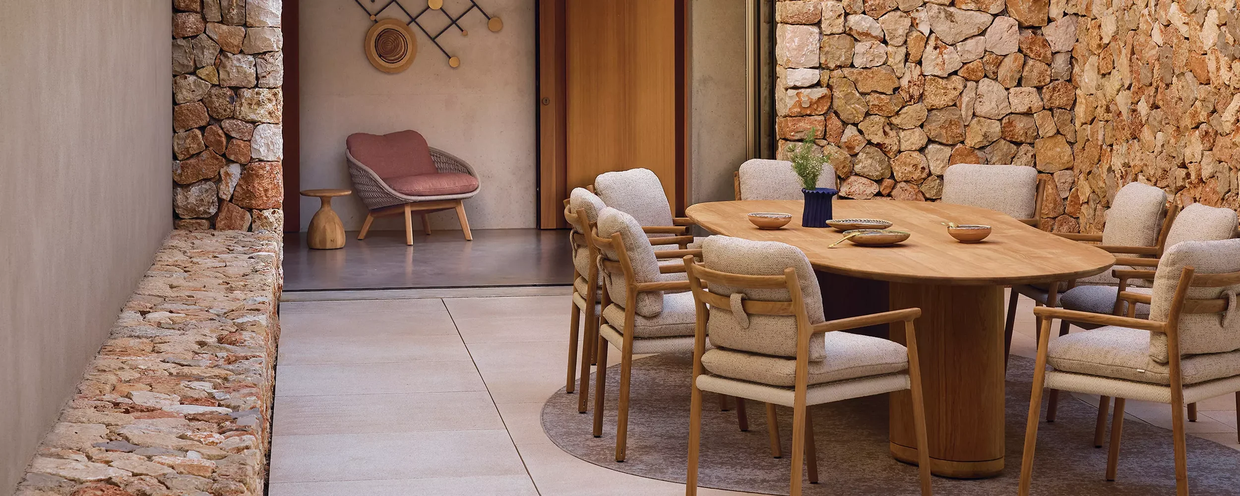 A dining room with a large wooden table surrounded by cream-colored upholstered chairs, a stone accent wall, and a small seating area with a pink cushion chair and a small wooden side table.