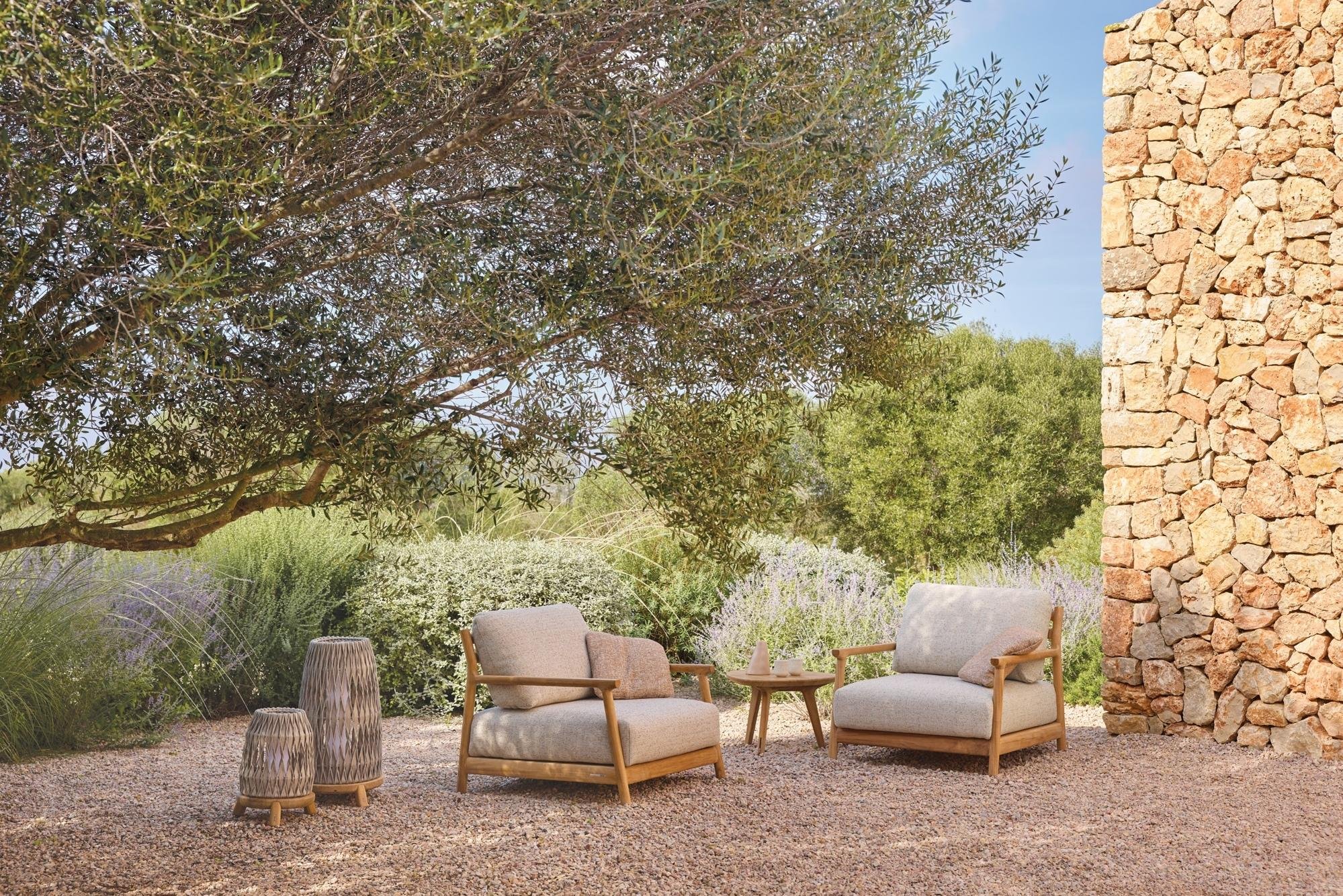 Outdoor patio with two beige cushioned armchairs with wooden frames, a small wooden side table, two decorative woven lanterns, surrounded by lush green trees and bushes, with a stone wall on the right and a gravel ground.