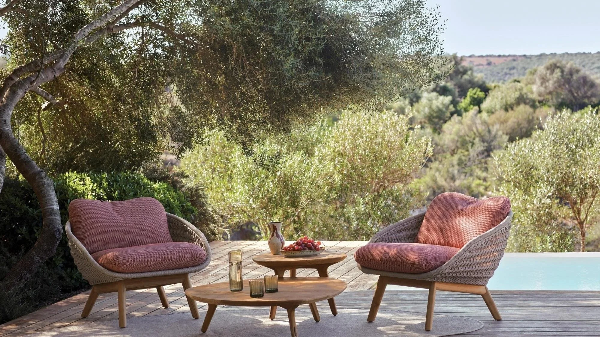 Outdoor patio with two wicker chairs with pink cushions, wooden coffee tables with candles, a vase, and a bowl of fruit, surrounded by trees and green foliage, overlooking a scenic hillside.