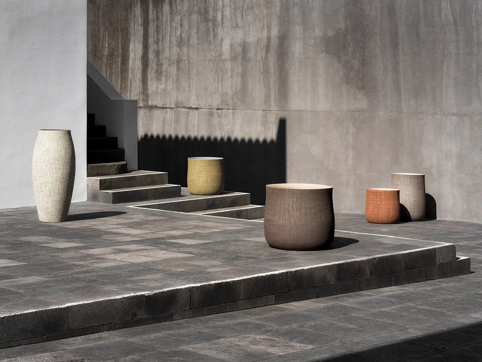 Five large textured ceramic planters in various pastel shades placed on a modern concrete patio with stairs and a plain concrete wall in the background.