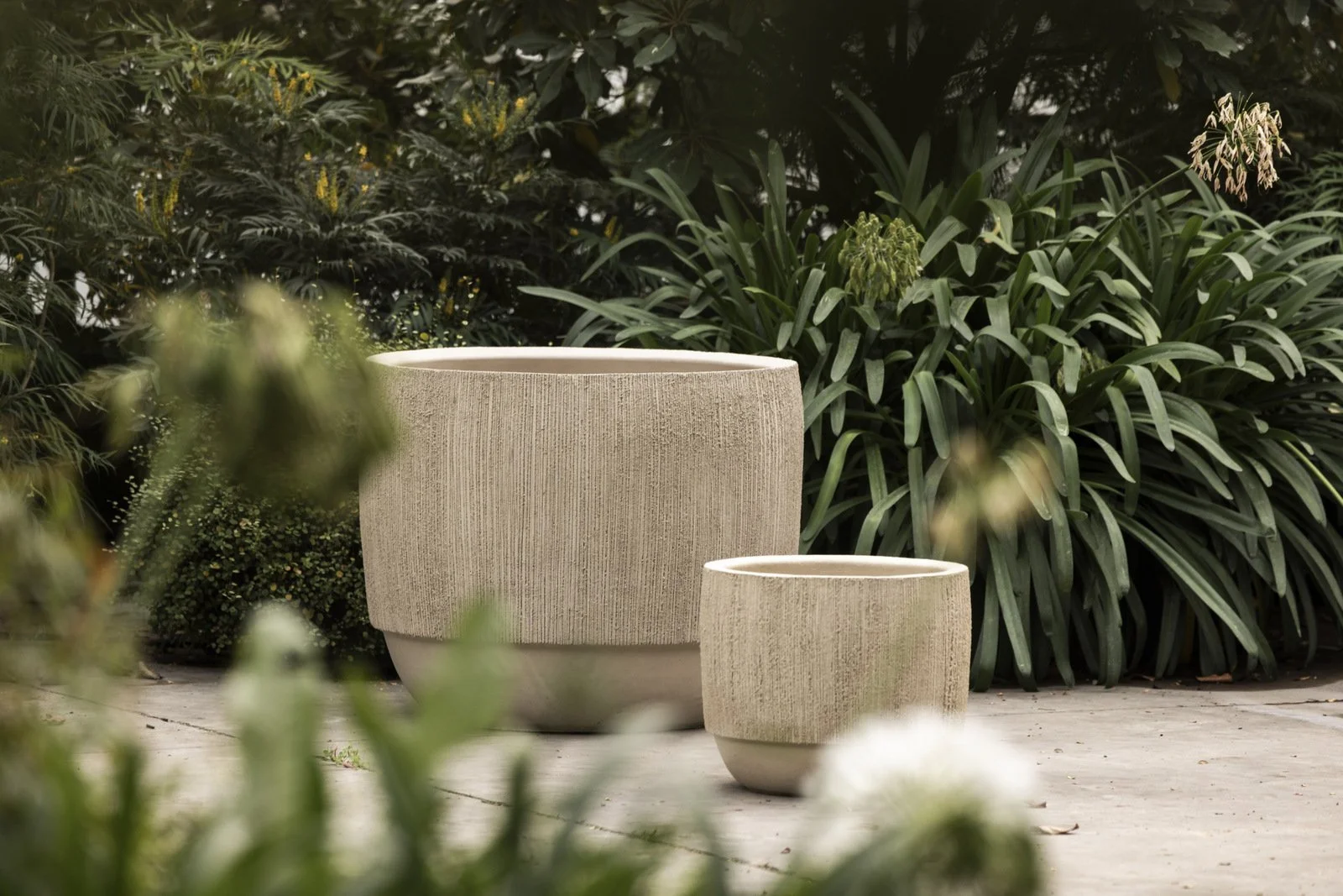 Two large beige textured planters with rounded shapes and a rough, ribbed surface, set outdoors on a patio with lush green foliage in the background.
