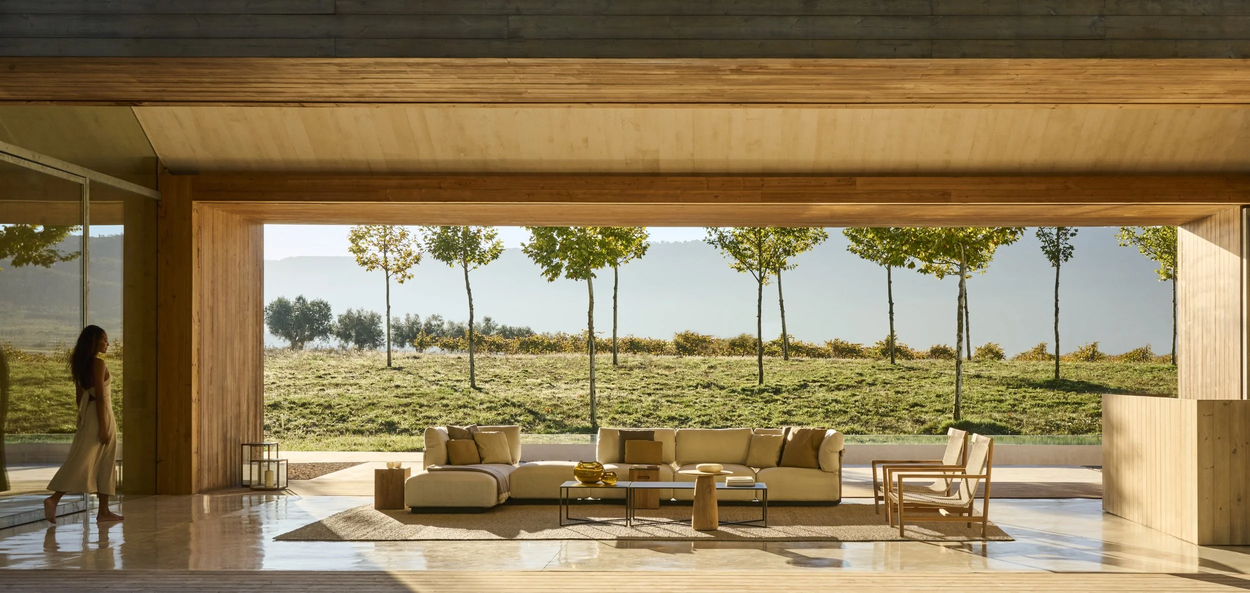 Modern living room with large open window overlooking a grassy yard with trees, beige sectional sofa, and wooden furniture.