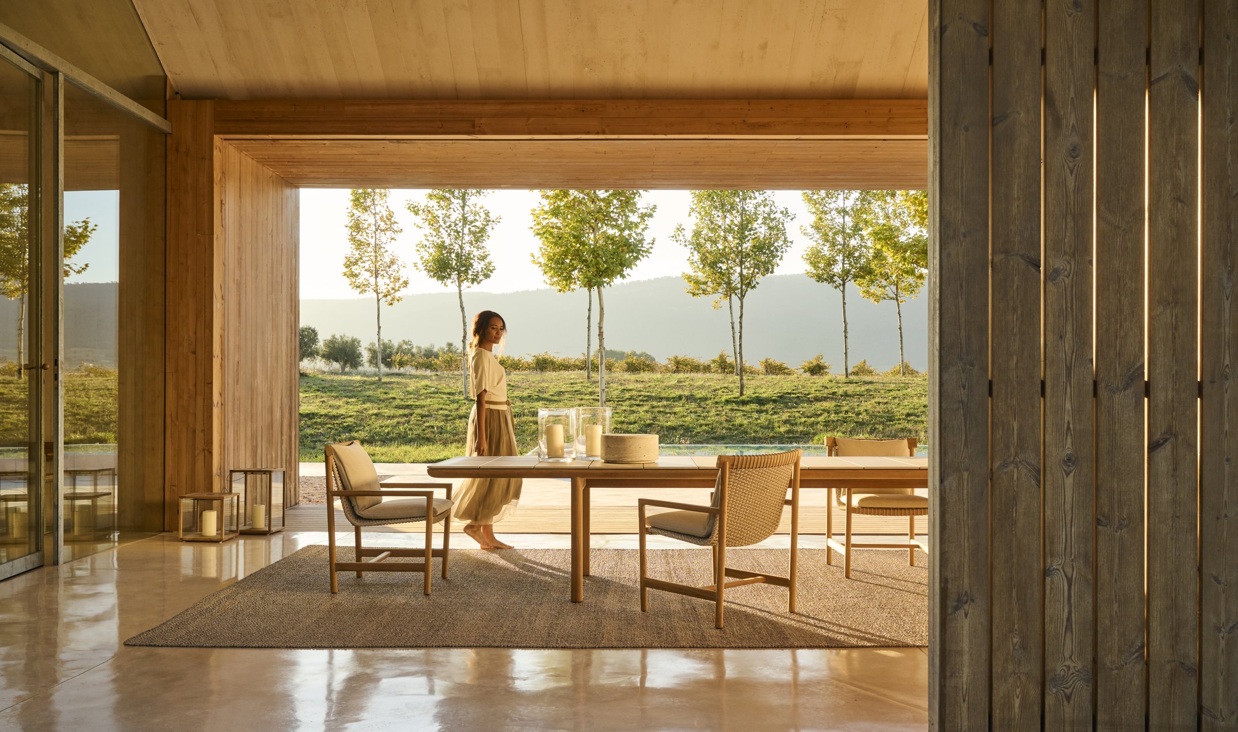 A woman standing on a patio outside a modern house, with trees and hills in the background during sunset.