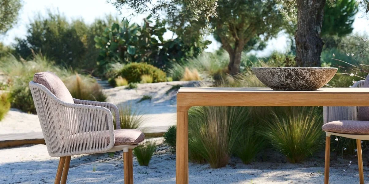 Outdoor scene with a wooden table, two chairs with cushions, and a large stone bowl, surrounded by trees and plants in a sandy area