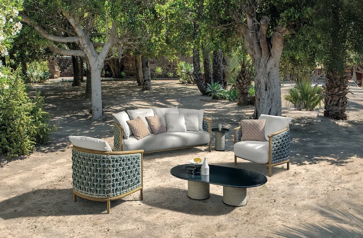 Outdoor seating area with a modern sofa and two chairs with patterned sides, a black coffee table, and small side tables, set in a sandy area with trees and plants in the background.