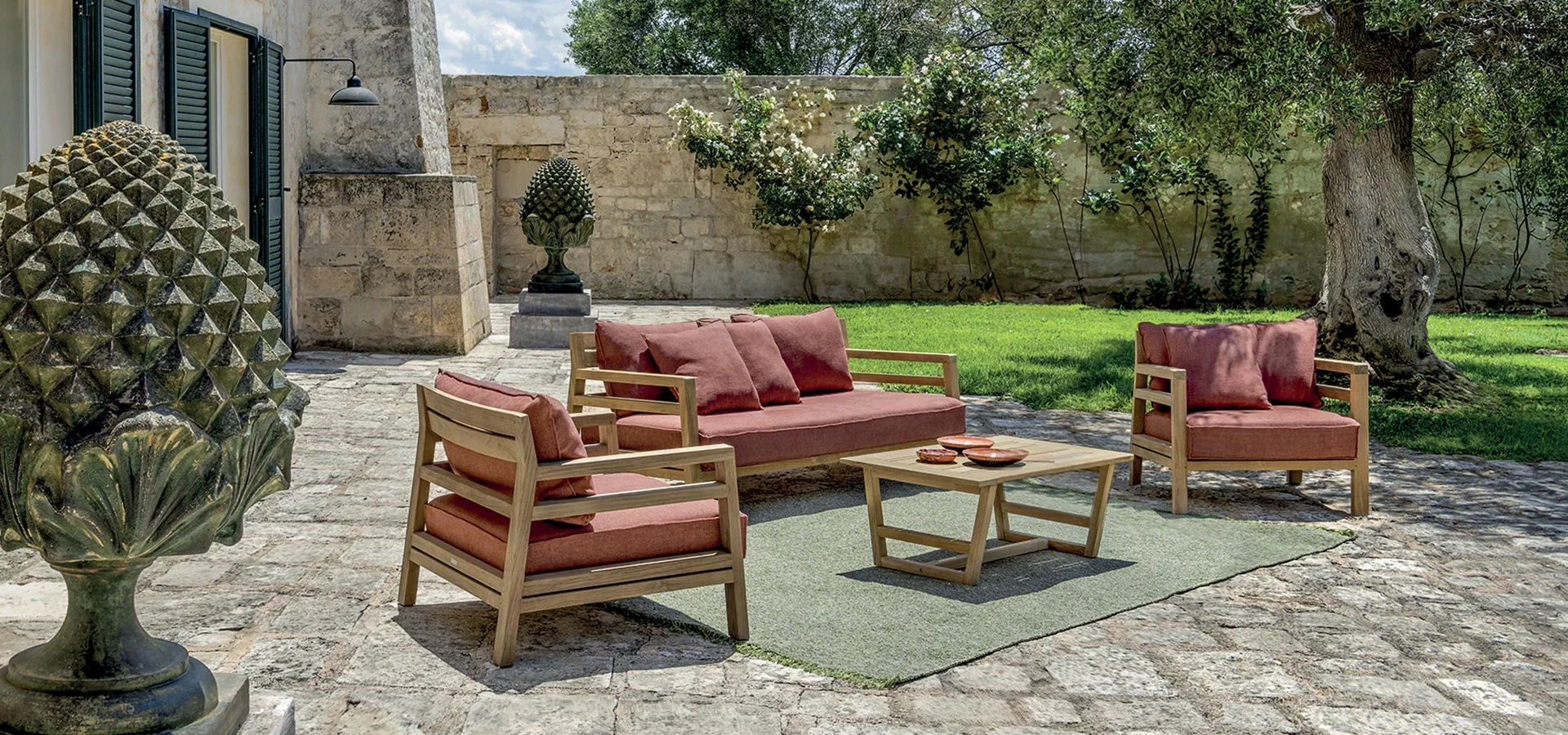 Outdoor patio with wooden furniture featuring pink cushions, a stone fountain, a stone statue, a green grassy area, large trees, and a stone wall in the background.