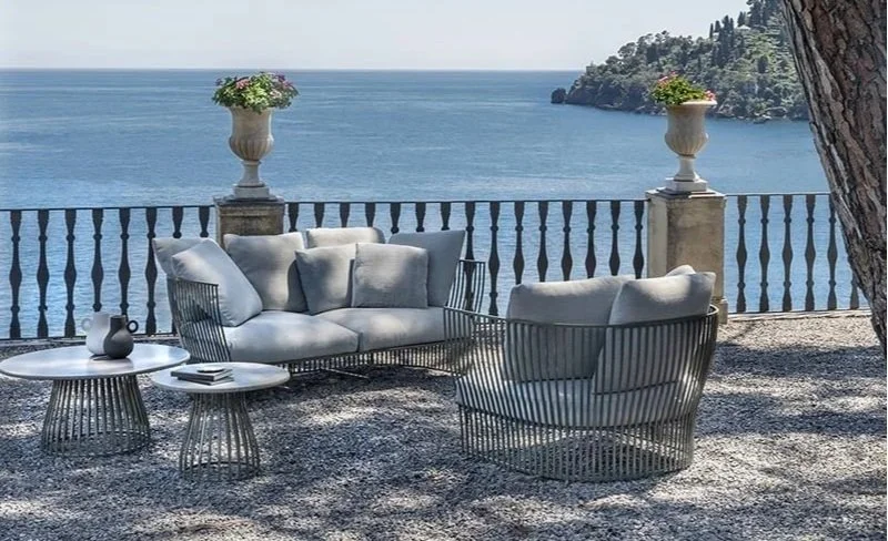 Outdoor seating area with a sofa, a chair, and two side tables overlooking a large body of water and a distant shoreline.