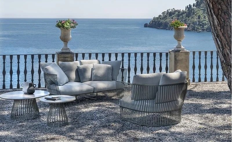 Outdoor seating area with a sofa, a chair, and two side tables overlooking a large body of water and a distant shoreline.