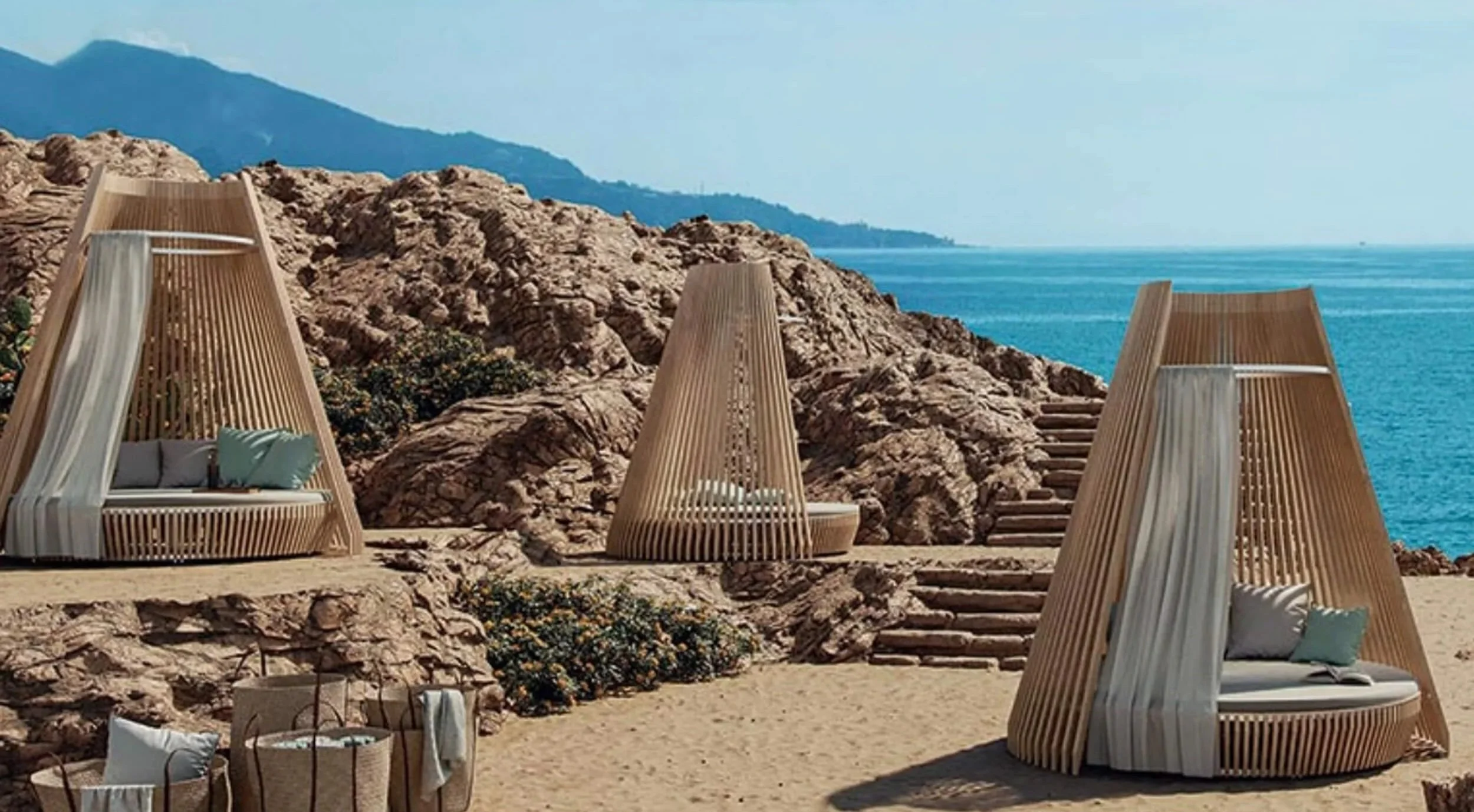 Beachside cabanas with wooden slats, cushions, and curtains on sandy ground with rocky backdrop and ocean in the background.