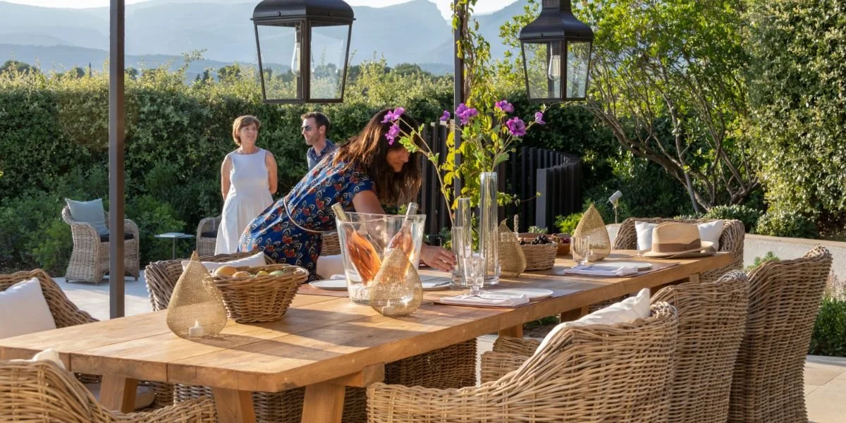 Outdoor dining table set with wicker chairs, glass vases, and linens, surrounded by green bushes and trees, with mountains in the background and two women and one man standing and one woman arranging items on the table.