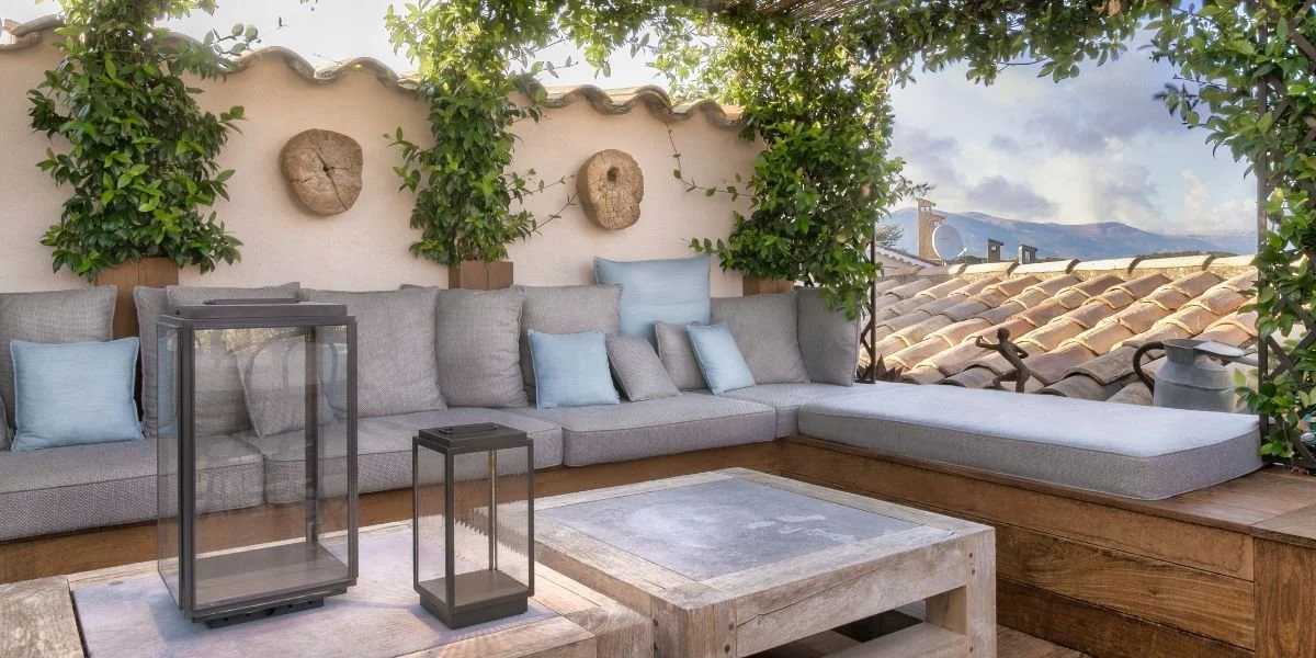 Outdoor patio with gray sectional sofa, glass lanterns, and wooden coffee table. Wall decorated with wooden art and surrounded by greenery.