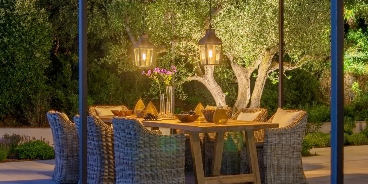 An outdoor dining area with a wooden table and wicker chairs, illuminated by hanging lanterns, surrounded by trees and greenery.
