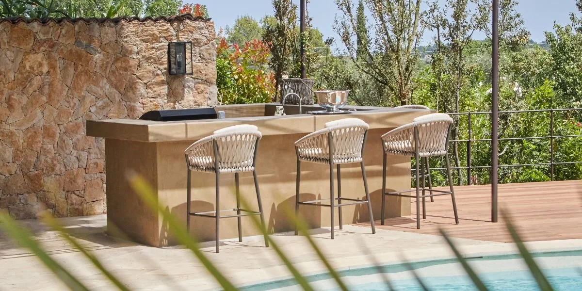 An outdoor bar area with a stone wall, four white wicker barstools, a beige bar counter, a sink, and a lush green garden background.