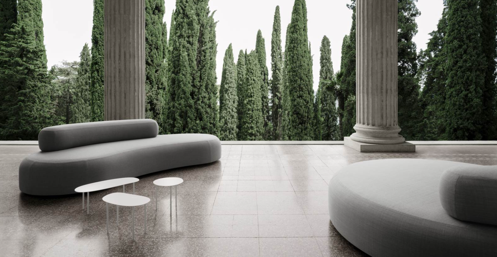 Modern outdoor patio with large stone columns, a curved grey couch, a rounded white lounge chair, and three small white tables, overlooking tall green cypress trees.