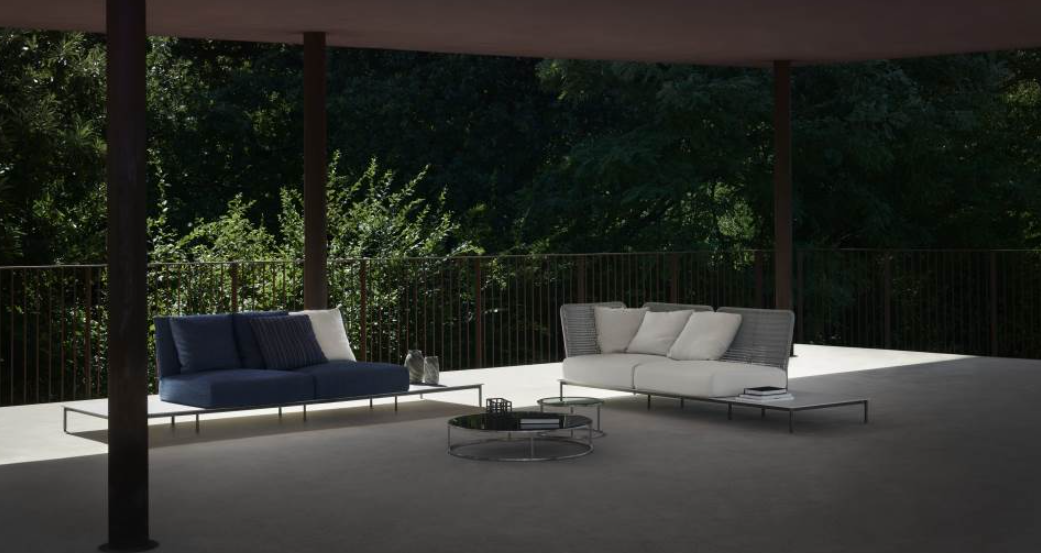 Outdoor patio with modern two-seat sofas, one blue with pillows and one white with pillows, and a round coffee table, overlooking a green wooded area.