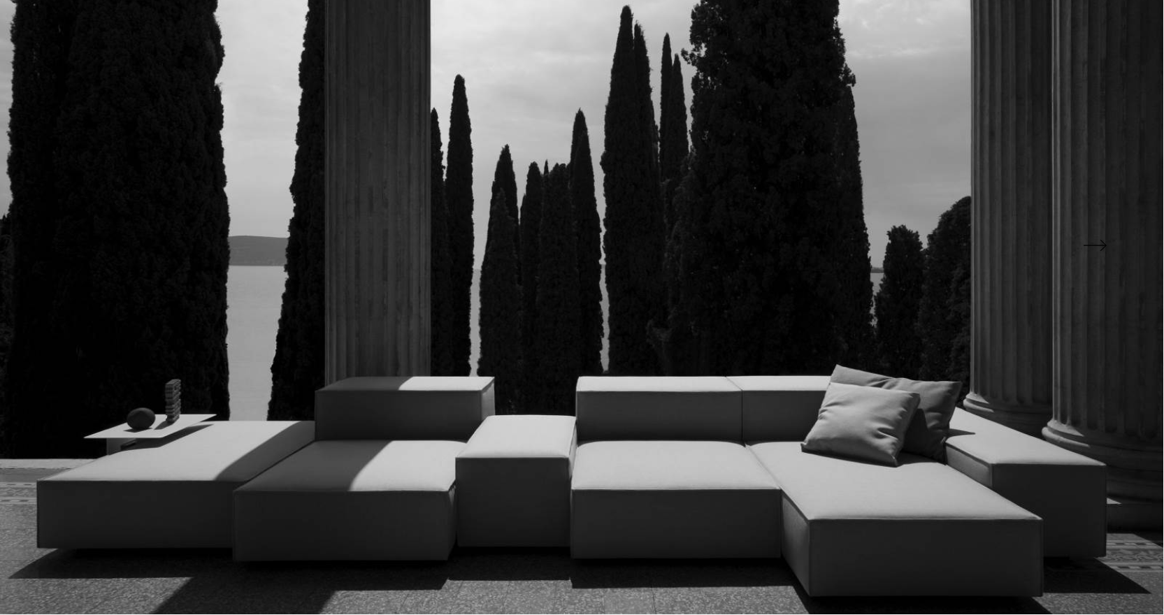 Outdoor patio with modern sectional sofa, pillows, side table with decorative objects, tall cypress trees and classical columns in the background, black and white photo.